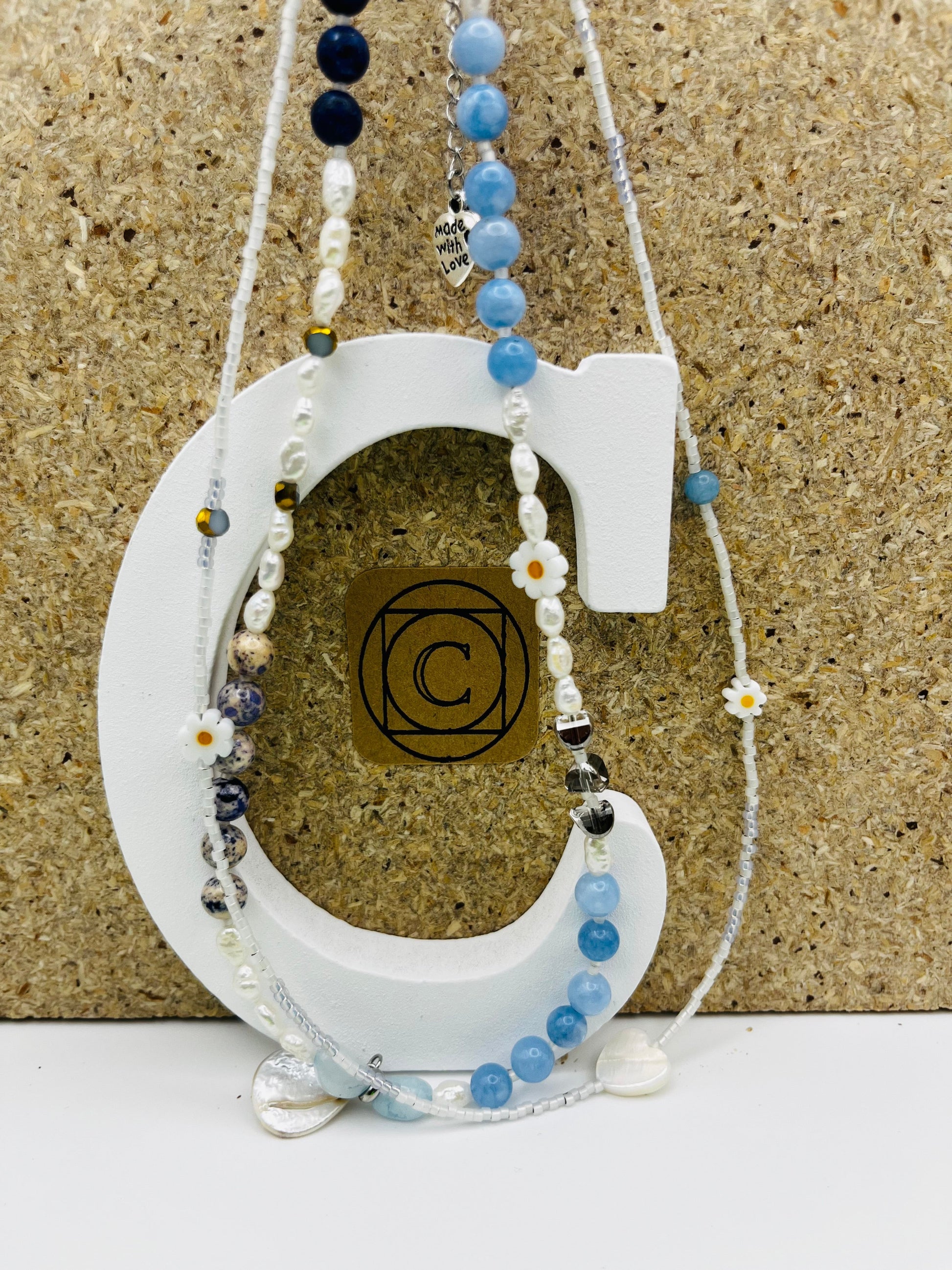 Collar Periwinkle – doble collar con semillas de rocalla blancas, margaritas de cerámica blancas con centro naranja, perlas barrocas blancas, cuentas de aguamarina azul cielo facetadas, jaspe dálmata, cuenta dorada, cuenta negra y corazón colgante de cristal transparente con charm "Made with Love", sobre letra C blanca con logo Cuadratura y fondo de corcho, hecho a mano en Murcia

