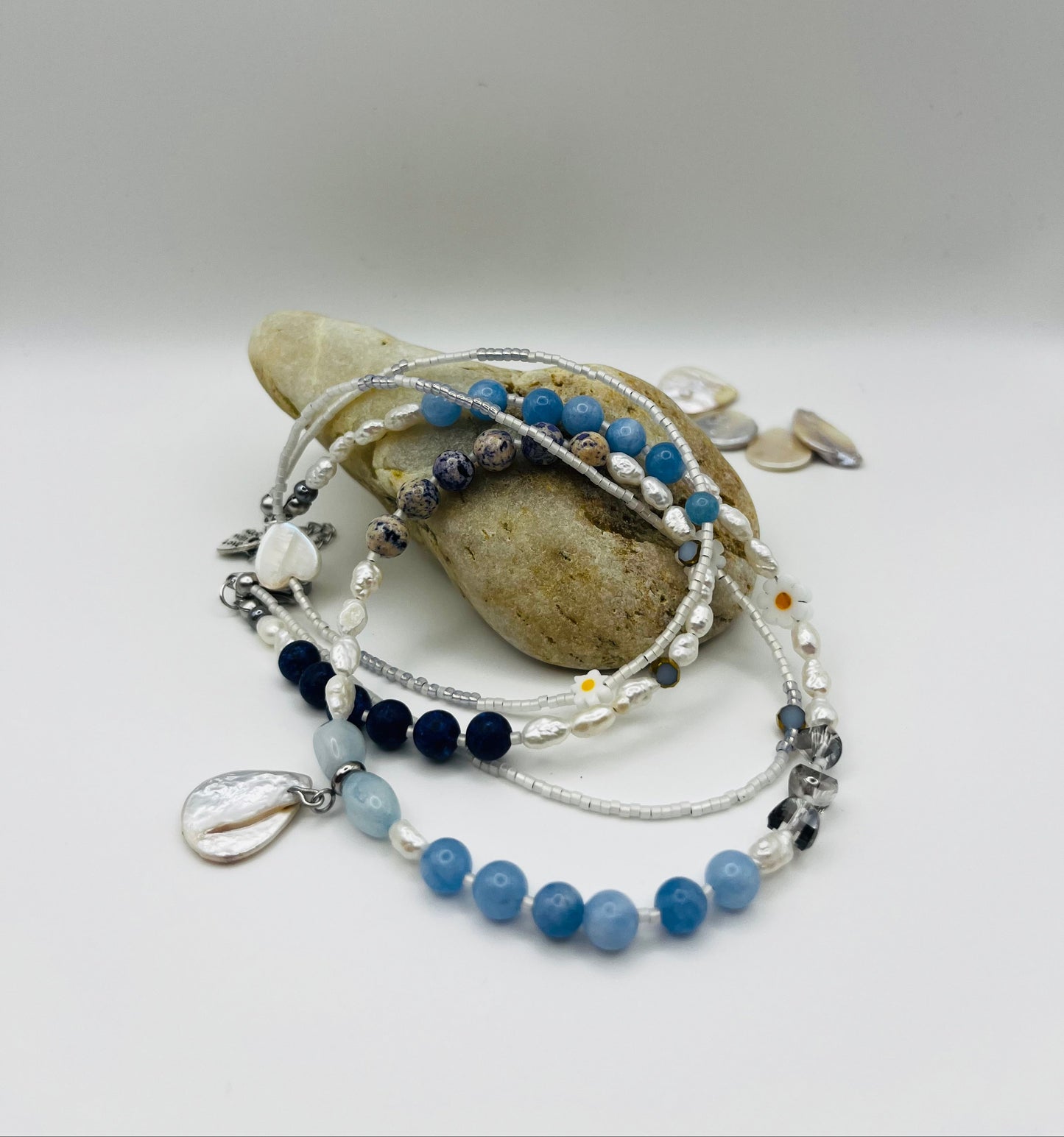 Collar Periwinkle – doble collar sobre piedra de playa con semillas de rocalla blancas, margaritas de cerámica blancas, perlas barrocas blancas, aguamarina azul cielo facetada, jaspe dálmata, cuenta negra y corazón colgante de cristal, con perlas barrocas en forma de corazón sueltas al fondo, hecho a mano en Murcia

