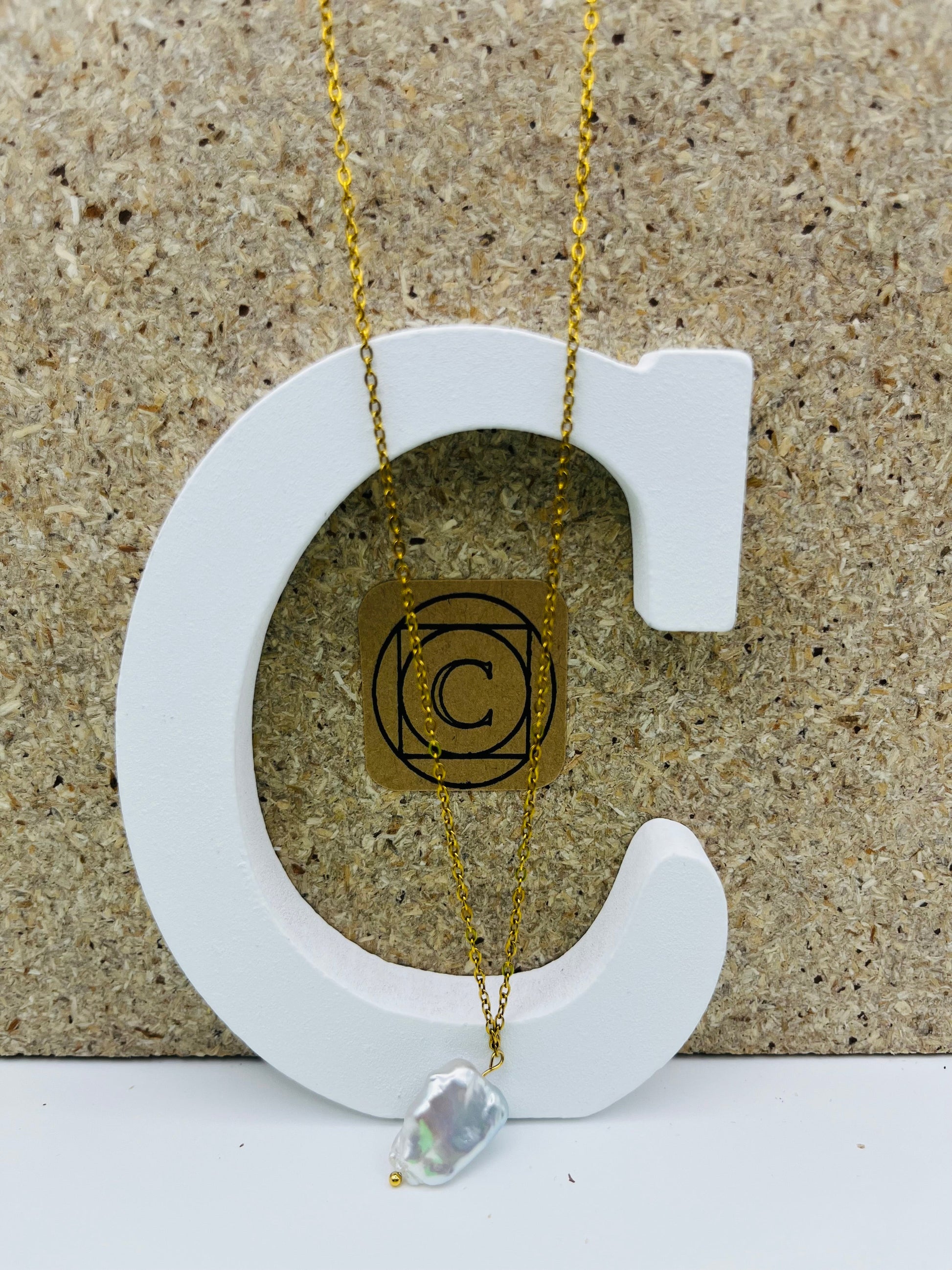 Collar minimalista con cadena dorada y perla barroca irregular blanca, presentado sobre letra C blanca con etiqueta dorada Cuadratura, montado a mano en Murcia
