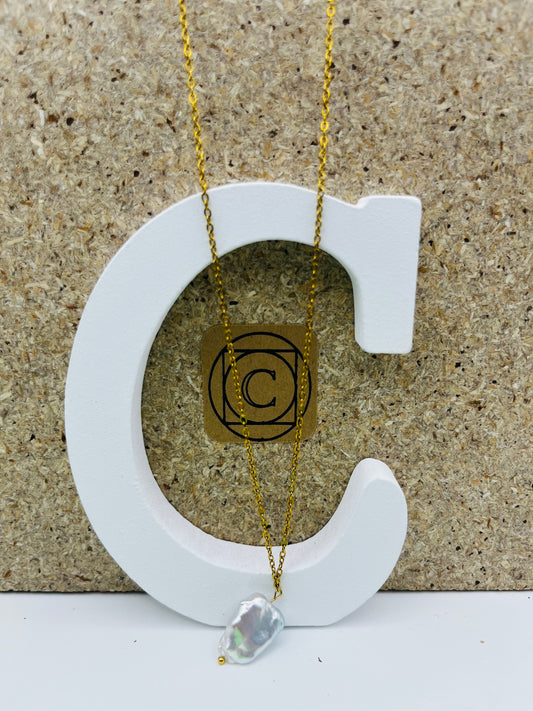 Collar minimalista con cadena dorada y perla barroca irregular blanca, presentado sobre letra C blanca con etiqueta dorada Cuadratura, montado a mano en Murcia
