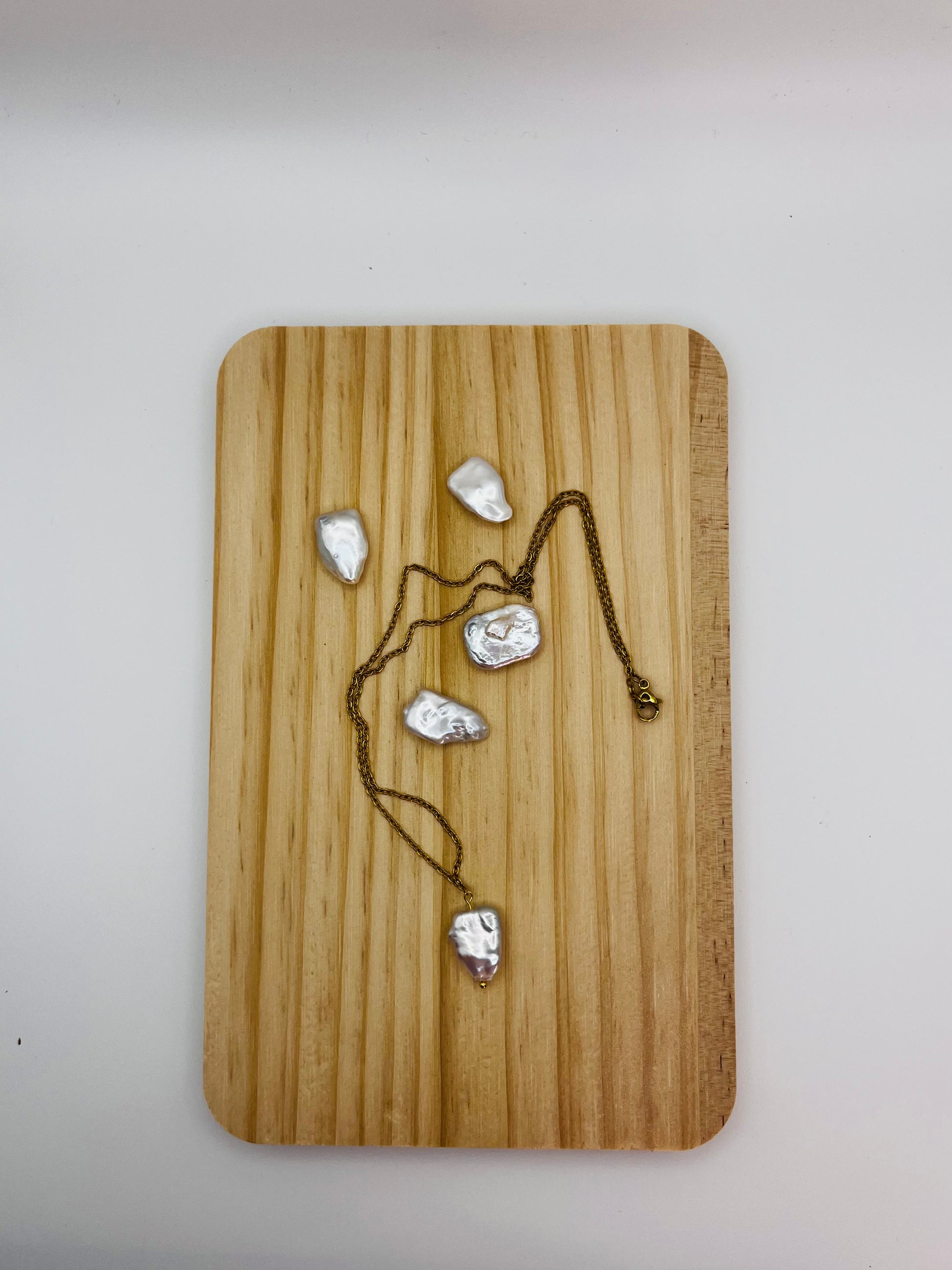 Collar minimalista con cadena dorada y perla barroca irregular blanca, sobre tabla de madera rodeado de perlas barrocas sueltas, montado a mano en Murcia

