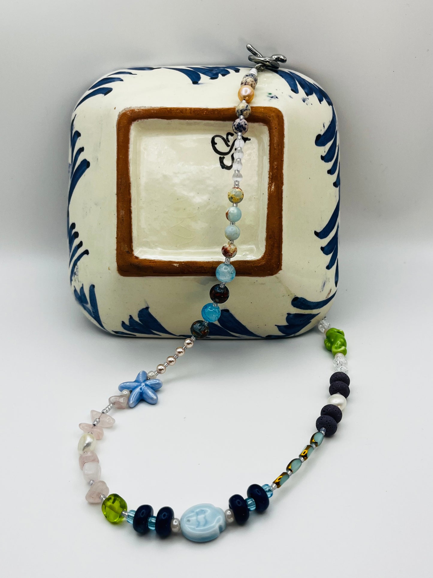Collar Salina – collar largo con cuentas de aguamarina, cuarzo rosa, lava volcánica negra, perlas barrocas, estrella de mar y pez de cerámica azul, caballito de mar verde y cierre toggle plateado, sobre azulejo cerámico azul y blanco de estilo mediterráneo, hecho a mano en Murcia