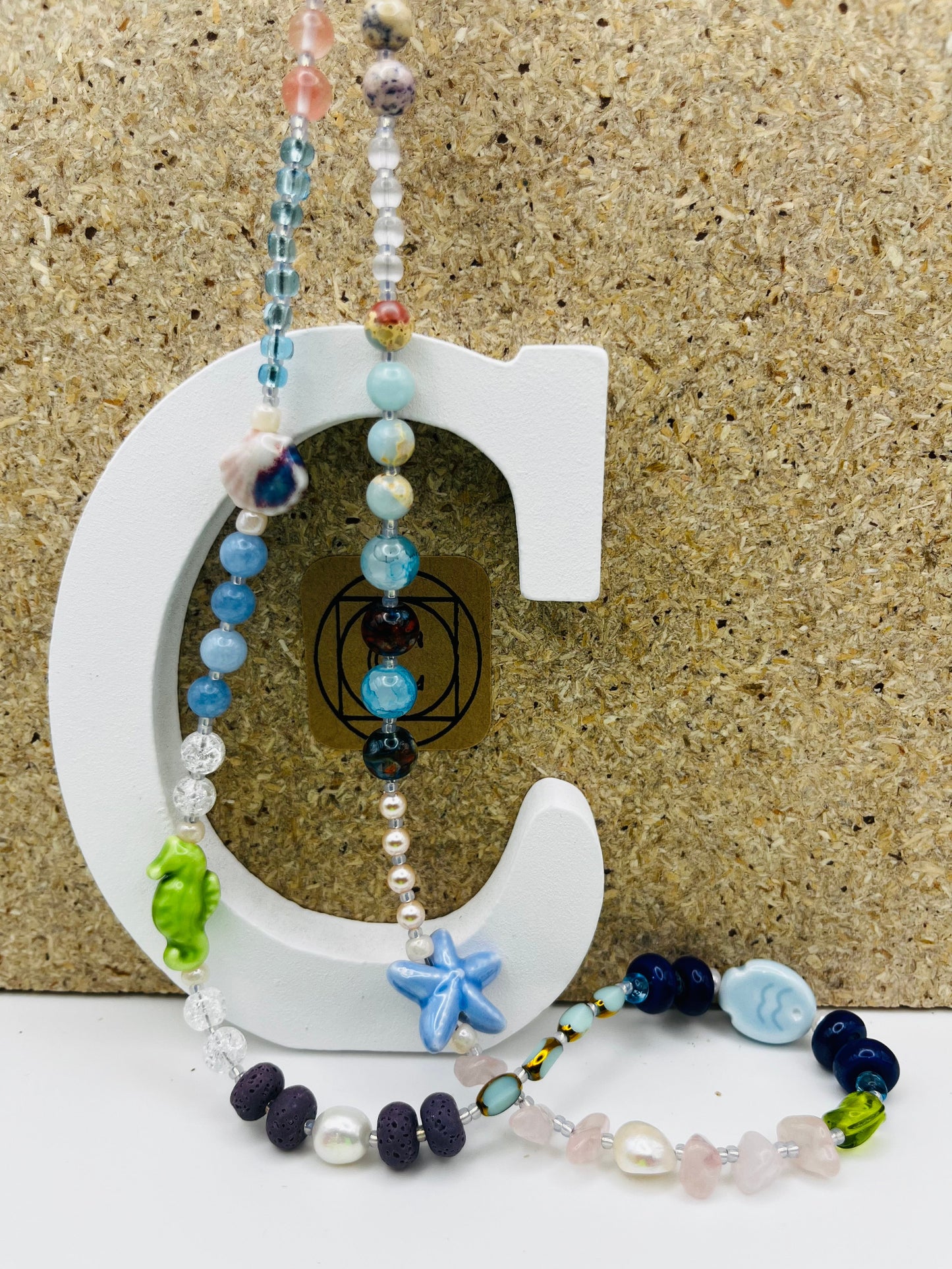 Collar Salina – collar largo con semillas de rocalla transparentes y azules, cuentas de cuarzo rosa, lava volcánica negra, perlas barrocas blancas, estrella de mar de cerámica azul, caballito de mar verde, cuentas de cristal azul y pez de cerámica azul, sobre letra C blanca con logo Cuadratura, hecho a mano en Murcia