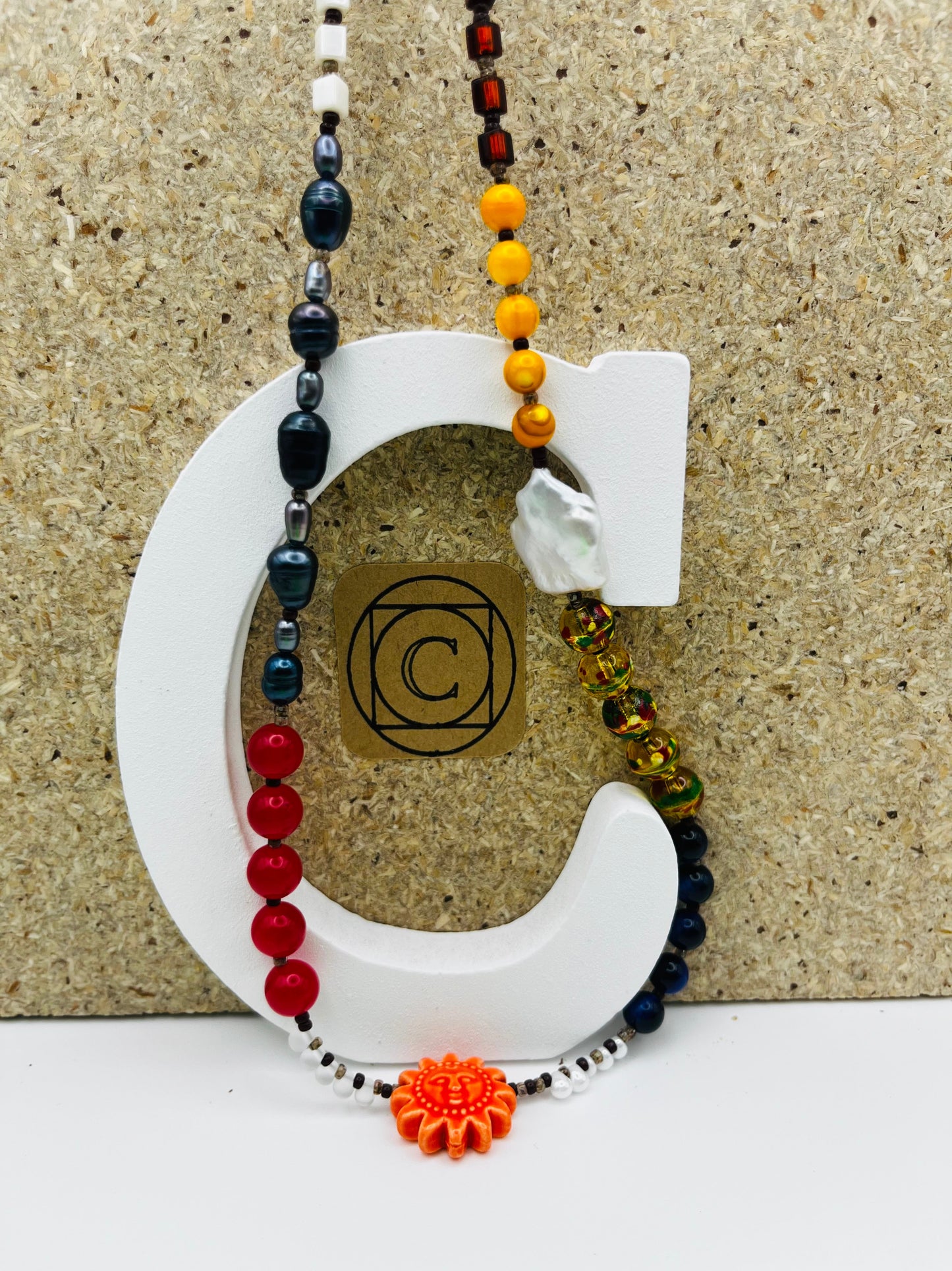 Collar Solana – collar largo con semillas de rocalla blancas y negras, perlas barrocas antracita, coral rojo, ágata, cloisonné multicolor, lapislázuli, cristal granate y sol de cerámica naranja, sobre letra C blanca con logo Cuadratura, hecho a mano en Murcia