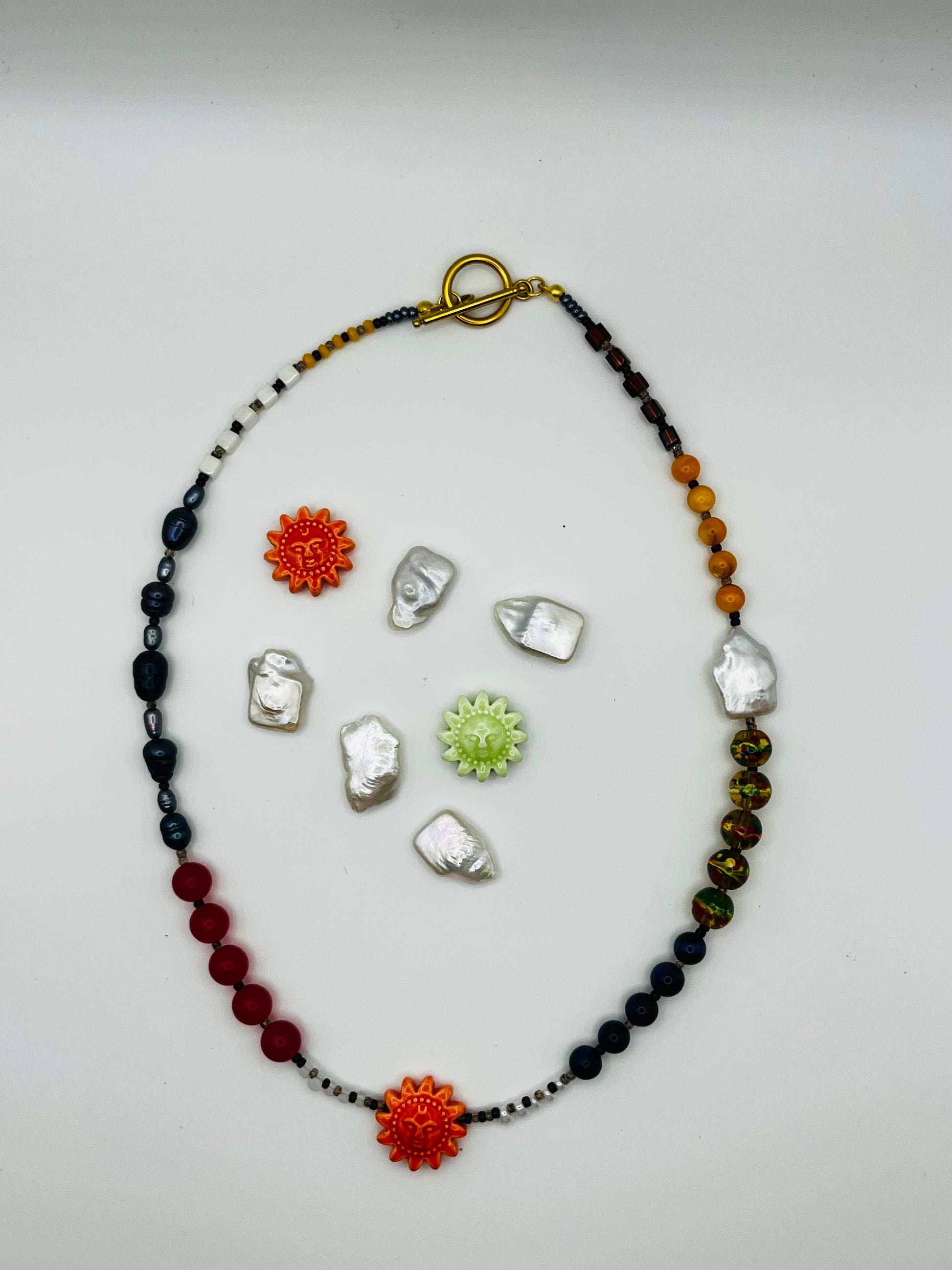 Collar Solana – collar largo con semillas de rocalla blancas y negras, perlas barrocas antracita, coral rojo, ágata, cloisonné multicolor, lapislázuli, cristal granate y sol de cerámica naranja, rodeado de soles de cerámica y perlas keshi sueltas, hecho a mano en Murcia
