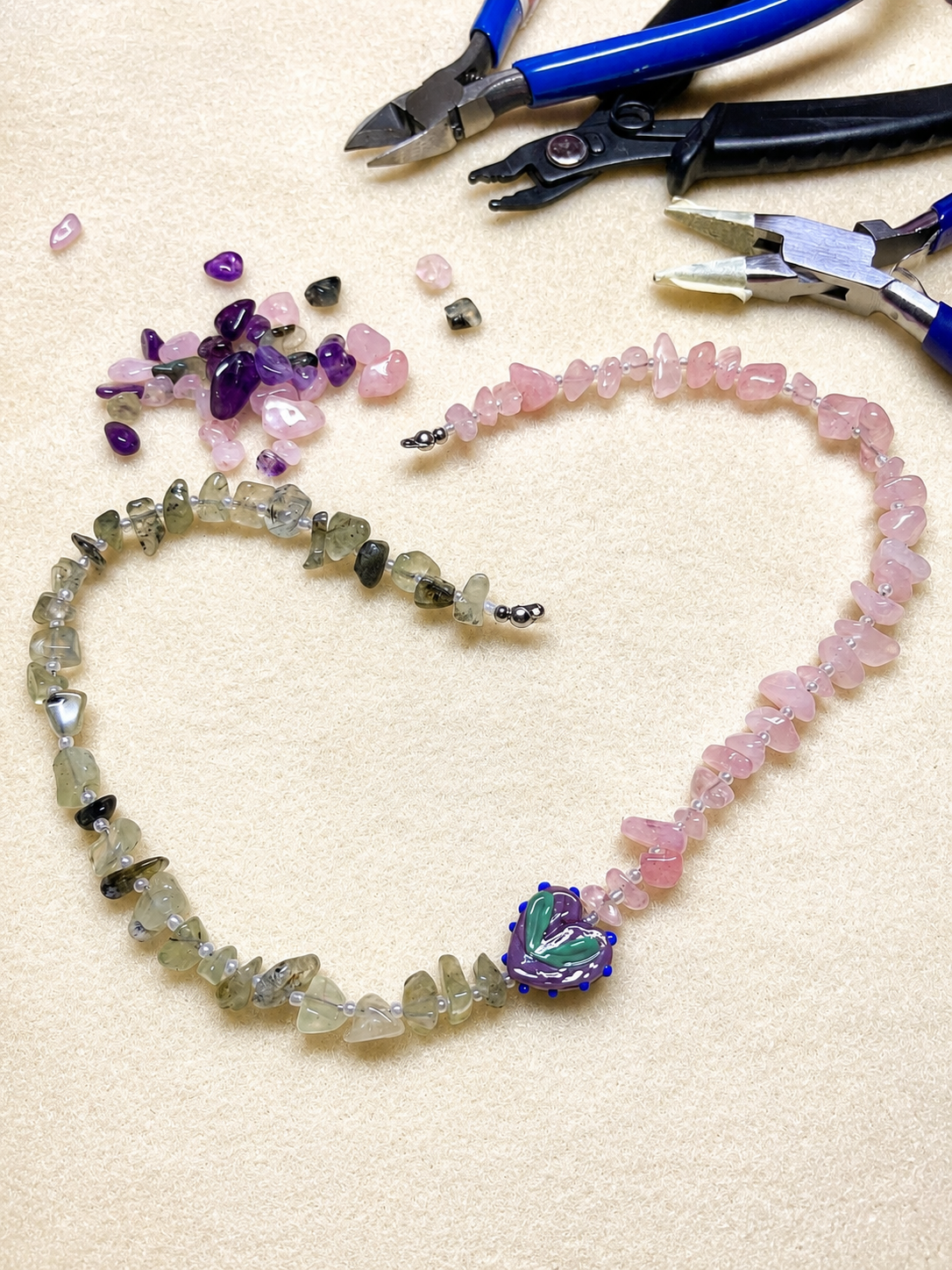 Collar Toulouse en proceso de elaboración artesanal, dispuesto en forma de corazón con chips de howlita, amatista y pieza central de vidrio soplado, junto a herramientas de joyería.