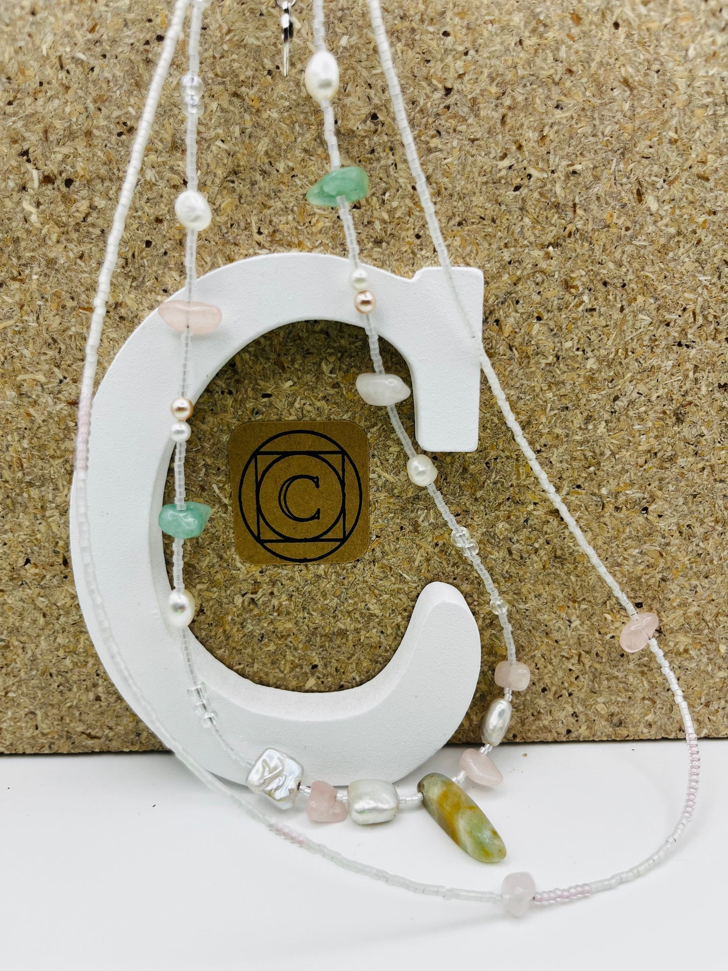 Collar Vera – doble collar con semillas de rocalla blancas y transparentes, perlas pequeñas de agua dulce, bolitas plateadas, chips de cuarzo rosa, chips de jade verde, perla barroca cuadrada blanca y cuenta alargada de jade verde oliva, sobre letra C blanca con logo Cuadratura y fondo de corcho, hecho a mano en Murcia