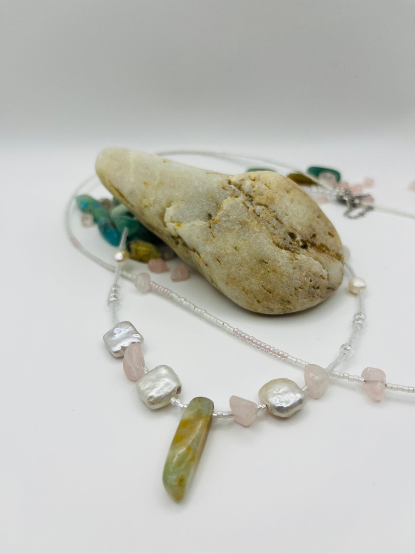 Collar Vera – doble collar con semillas de rocalla blancas y transparentes, perlas barrocas cuadradas blancas, chips de cuarzo rosa, bolitas plateadas y cuenta alargada de jade verde oliva, apoyado sobre piedra de playa con chips de jade y cuarzo rosa sueltos, hecho a mano en Murcia