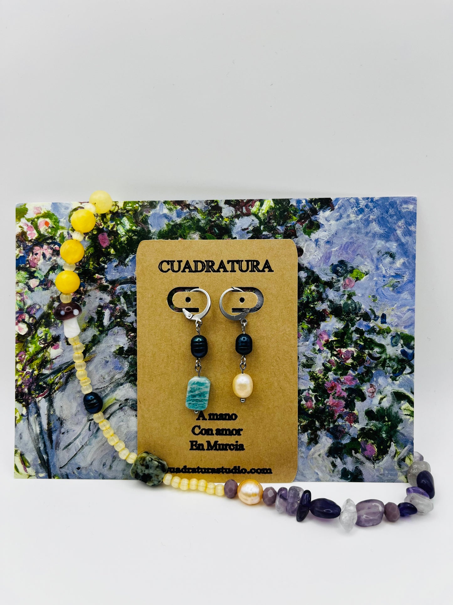 Conjunto Dalia – collar doble con jade amarillo, amatista, cuarzo, seta de cristal morada y charm de lazo plateado, junto a Pendientes Dalia asimétricos con perla negra, amazonita verde y perla barroca blanca en tarjeta Cuadratura, sobre postal de pintura impresionista, hechos a mano en Murcia