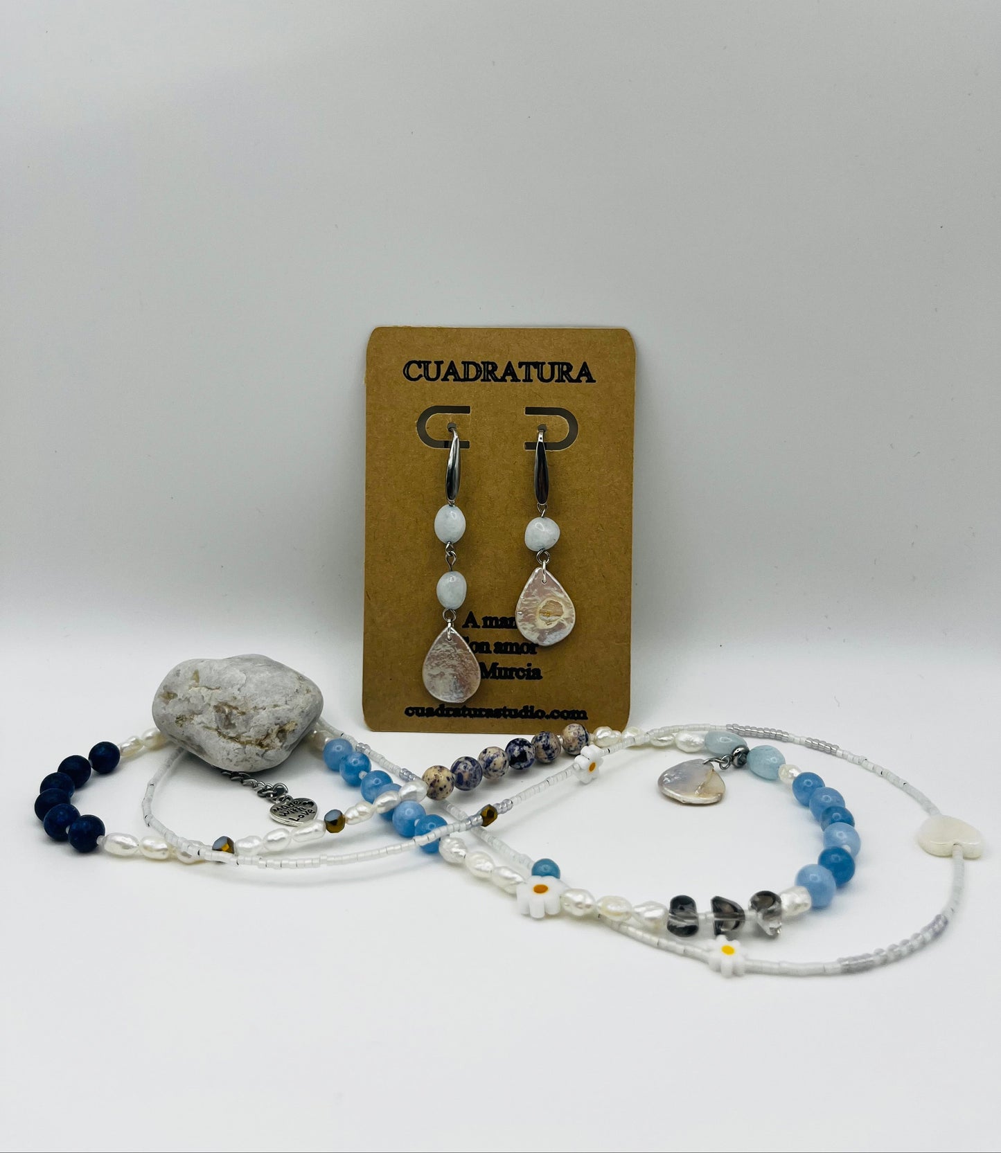 Conjunto Periwinkle – collar doble con semillas de rocalla blancas, margaritas de cerámica blancas, perlas barrocas blancas, aguamarina azul cielo facetada, jaspe dálmata, cuenta negra y corazón colgante de cristal, junto a Pendientes Periwinkle con aro plateado, aguamarina y perla barroca en forma de lágrima, en tarjeta Cuadratura, con piedra de playa, hechos a mano en Murcia