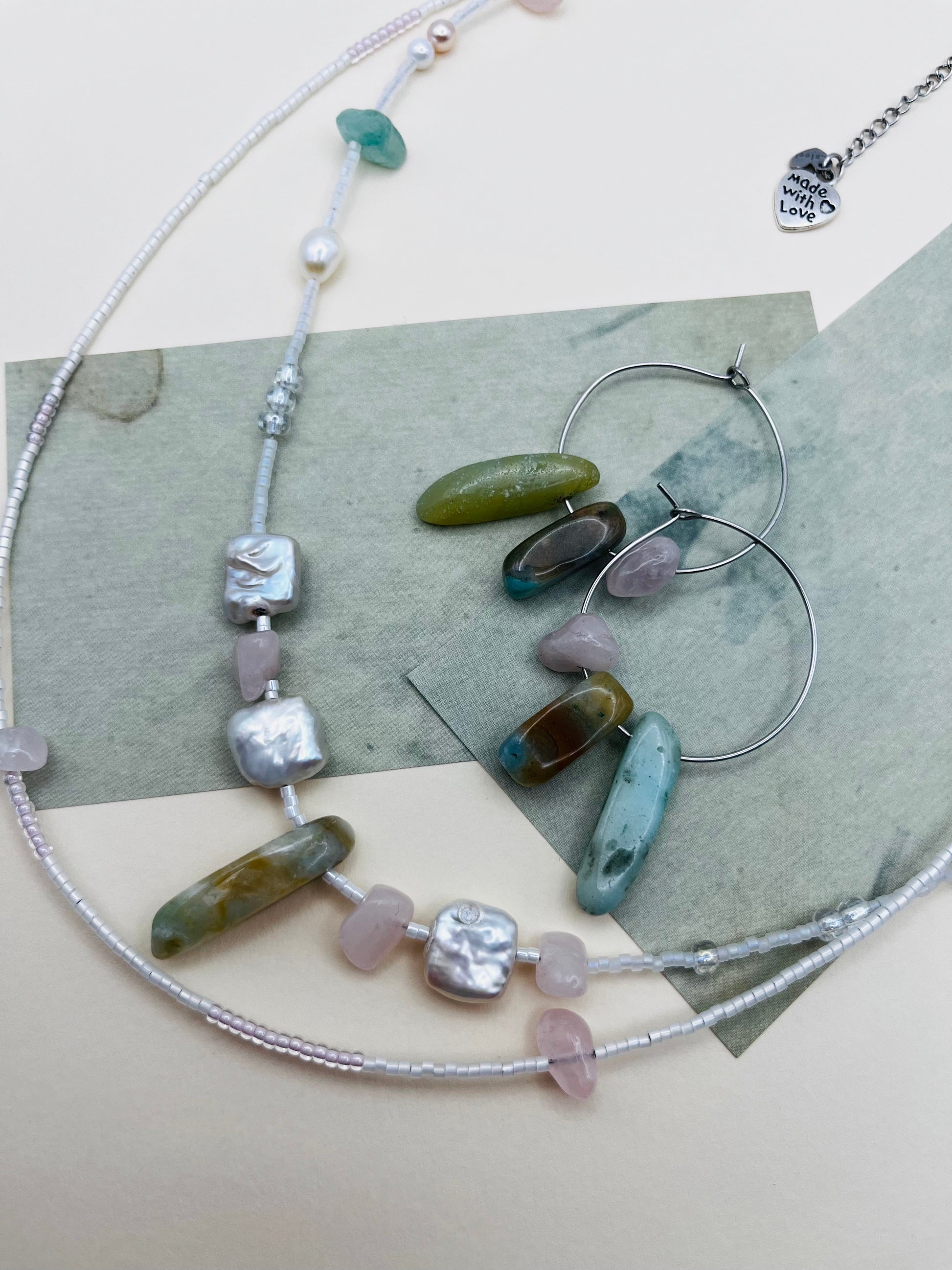 Conjunto Vera – collar doble con semillas de rocalla blancas, perlas barrocas cuadradas, chips de cuarzo rosa, jade verde y cuenta alargada de jade verde oliva, junto a Pendientes Levante con aro plateado y chips de jade verde y azul, sobre papel de acuarela verde grisáceo, hechos a mano en Murcia

