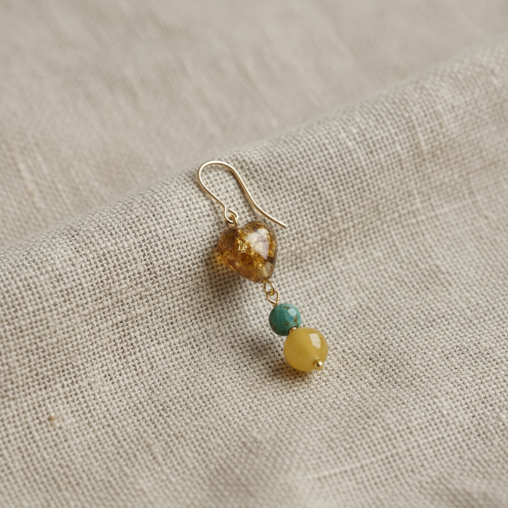 Pendiente Miel individual hecho a mano con corazón de cristal dorado, piedra verde y cuenta amarilla, sobre fondo de lino.