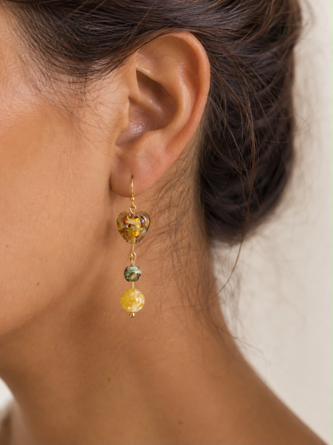 Mujer luciendo un pendiente Miel hecho a mano con corazón de cristal dorado, piedra verde y cuenta amarilla.