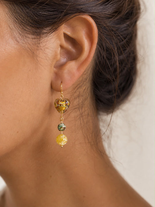 Mujer luciendo un pendiente Miel hecho a mano con corazón de cristal dorado, piedra verde y cuenta amarilla.