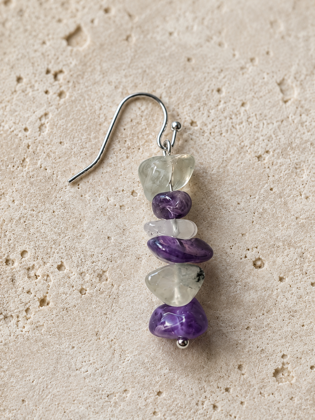 Detalle de un pendiente Verena con amatista y fluorita natural en tonos violeta, blanco y verde, montado en acero inoxidable sobre fondo de piedra.