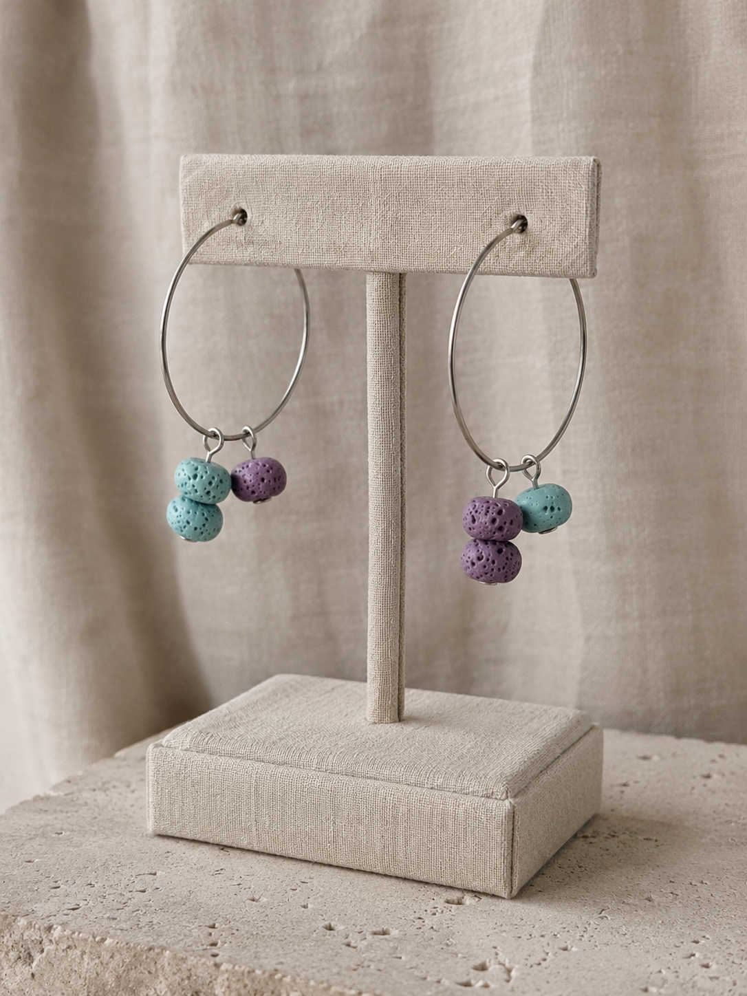 Pendientes artesanales Arrecife sobre expositor de joyería. Aros de acero inoxidable plateado con cuentas de piedra volcánica porosa en tonos turquesa y lila, en diseño asimétrico, sobre fondo de lino neutro.