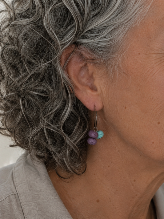 Pendientes artesanales Arrecife sobre modelo de pelo canoso rizado. Aros de acero inoxidable plateado con cuentas de piedra volcánica porosa en tonos turquesa y lila, en diseño asimétrico.