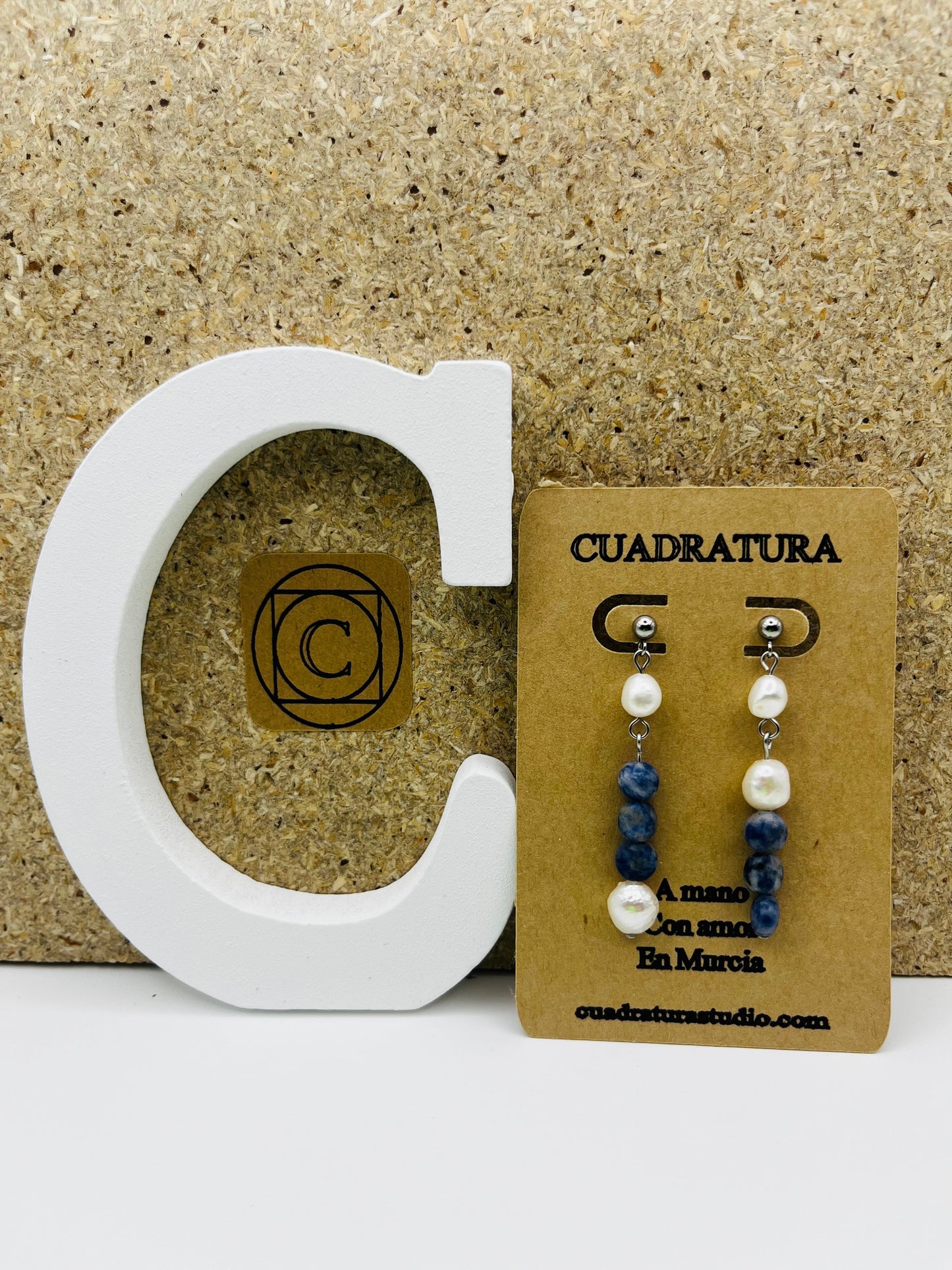 Pendientes Bajamar – pendientes simétricos con enganche plateado de bolita, perla oval blanca, chip de sodalita azul y perla oval blanca colgando, en tarjeta Cuadratura apoyada en letra C blanca, fondo de corcho, hechos a mano en Murcia