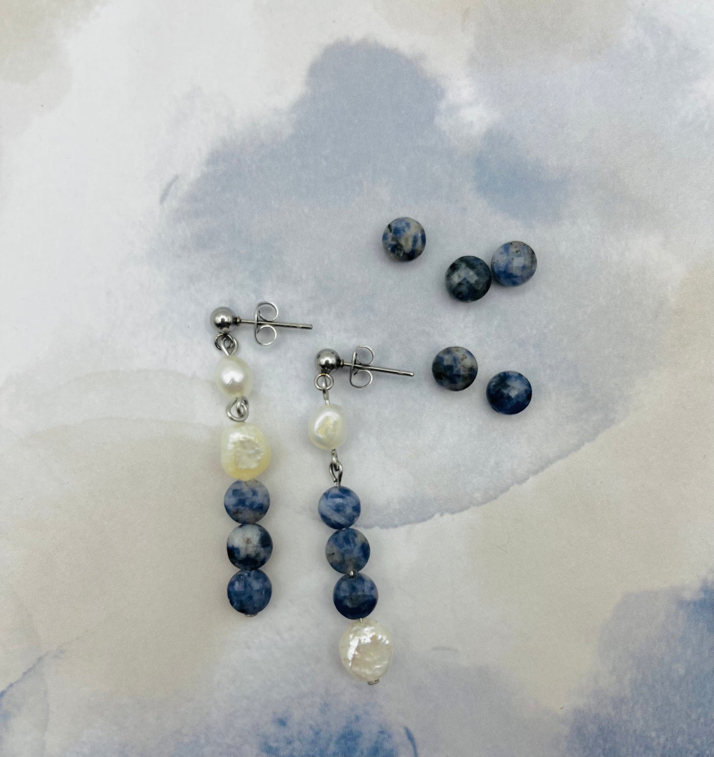 Pendientes Bajamar – pendientes simétricos con enganche plateado de bolita, perla oval blanca, tres cuentas redondas de sodalita azul y perla oval blanca en la base, desplegados sobre papel de acuarela azul grisáceo, con cuentas de sodalita redonda sueltas, fondo blanco, hechos a mano en Murcia