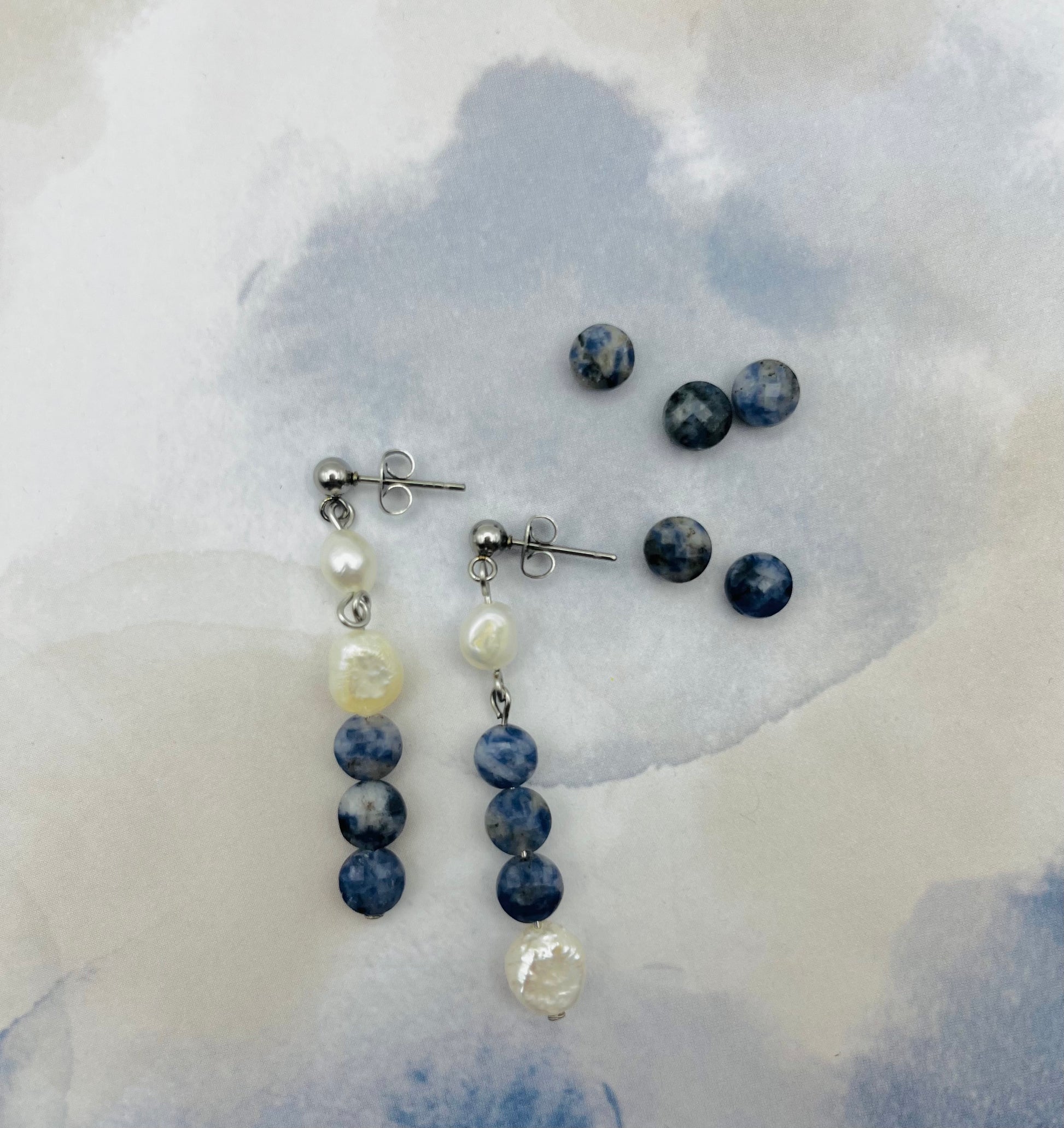 Pendientes Bajamar – pendientes simétricos con enganche plateado de bolita, perla oval blanca, tres cuentas redondas de sodalita azul y perla oval blanca en la base, desplegados sobre papel de acuarela azul grisáceo, con cuentas de sodalita redonda sueltas, fondo blanco, hechos a mano en Murcia