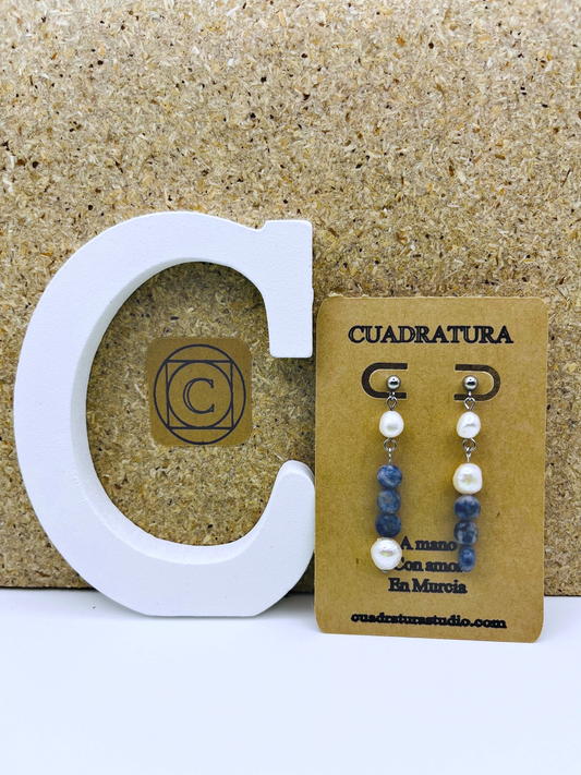 Pendientes Bajamar de Cuadratura Studio en tarjeta kraft: botón plateado con perla barroca blanca y tiras de sodalita azul, y botón con perla barroca y sodalita, sobre fondo de corcho con letra C blanca y logo de Cuadratura. Montados a mano en Murcia.`