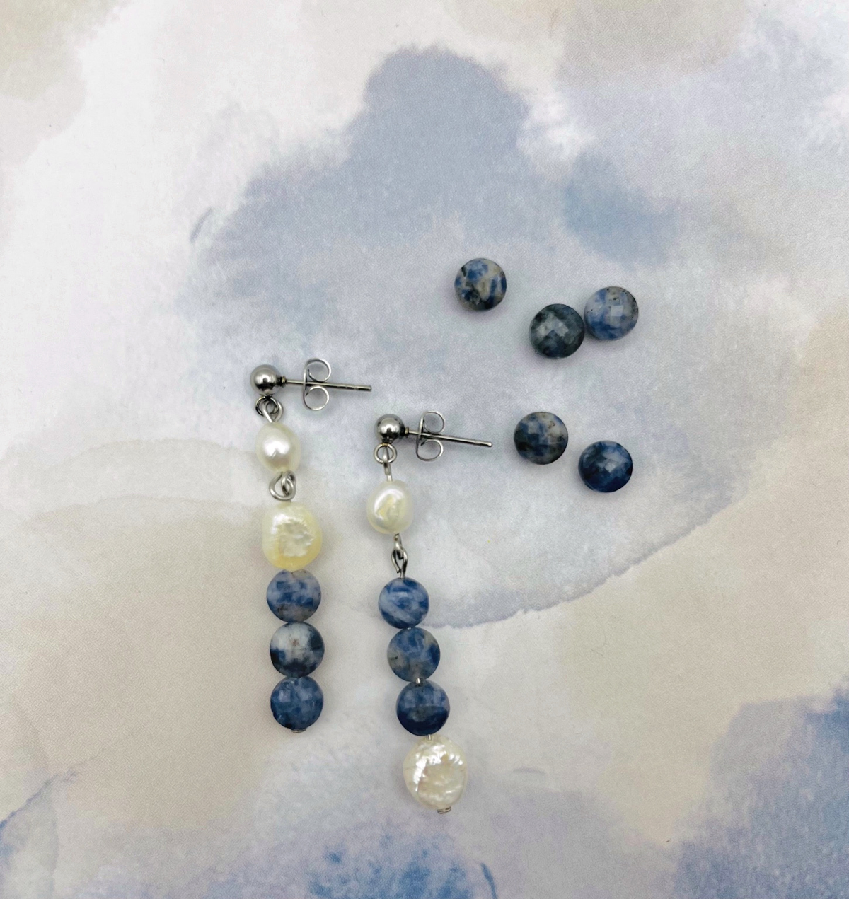 Pendientes Bajamar de Cuadratura Studio sobre fondo acuarela azul: pendiente largo asimétrico con botón plateado, perlas barrocas blancas y tres cuentas de sodalita azul mateada, junto a pendientes de tuerca de sodalita azul. Montados a mano en Murcia.`