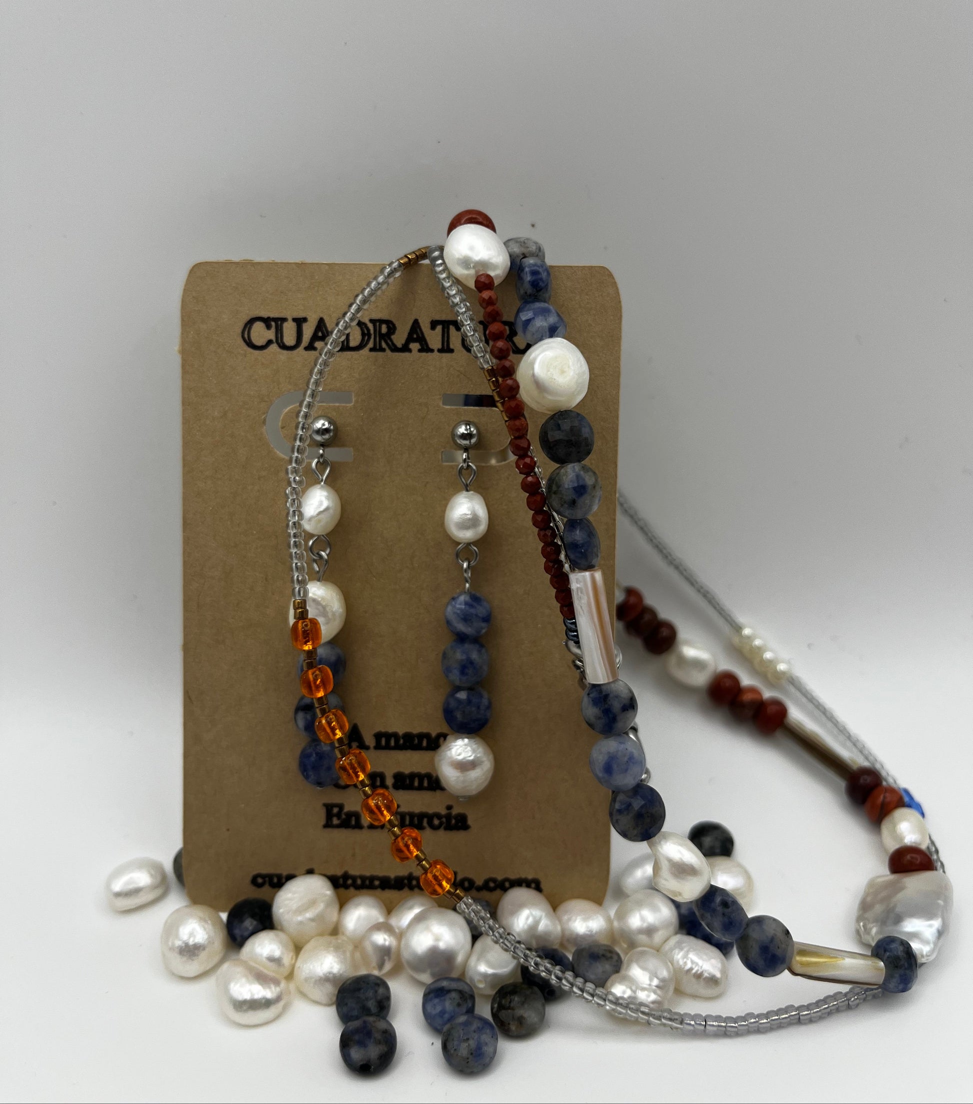 Pendientes Bajamar – pendientes simétricos con enganche plateado de bolita, perla oval blanca, tres cuentas de sodalita azul y perla oval blanca en la base, en tarjeta Cuadratura rodeada de collares de perlas oval blancas, sodalita azul y cornalina roja, fondo blanco, hechos a mano en Murcia