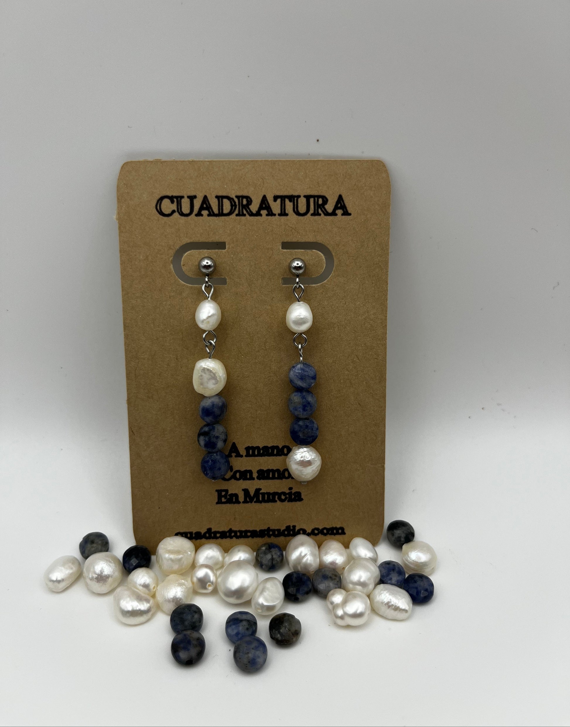 Pendientes Bajamar – pendientes simétricos con enganche plateado de bolita, perla oval blanca, tres cuentas redondas de sodalita azul y perla oval blanca en la base, en tarjeta Cuadratura, fondo blanco, con perlas oval blancas y cuentas de sodalita azul sueltas en la base, hechos a mano en Murcia