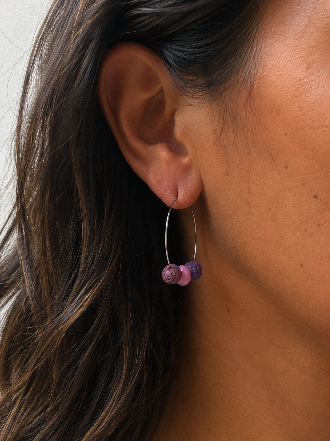 Mujer luciendo un pendiente Berry de aro en acero inoxidable con cuentas de piedra volcánica morada y cuentas de cristal rosa, hecho a mano.