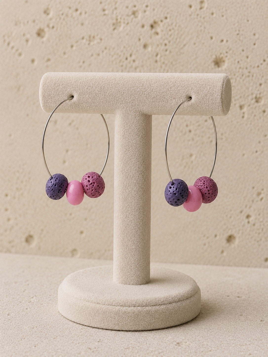 Pendientes Berry en expositor, aro de acero inoxidable con cuentas de piedra volcánica en tonos morado y rosa.