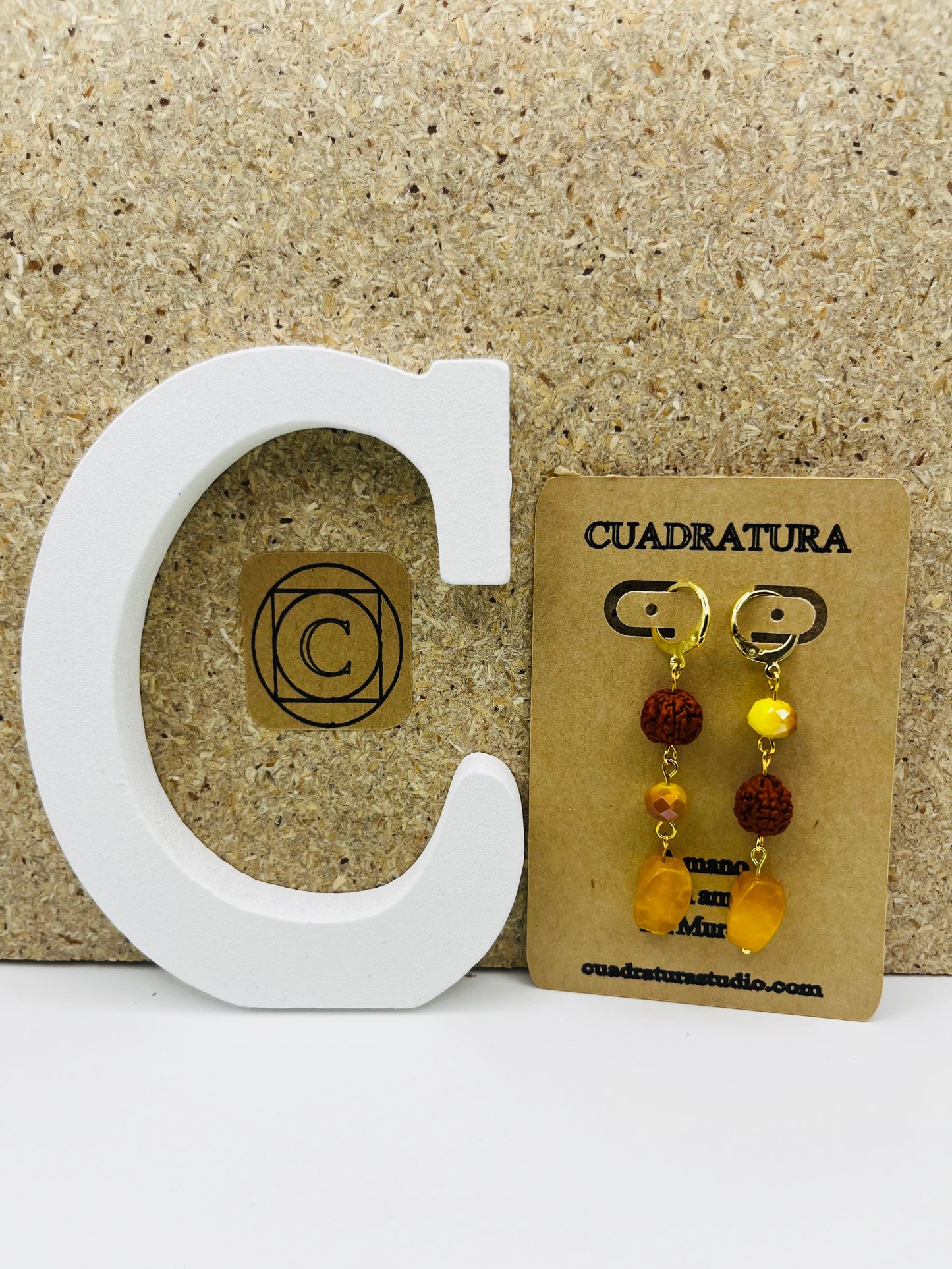 Pendientes Calima – pendientes simétricos con aro dorado, cuenta redonda de rudraksha marrón, cuenta facetada amarilla ámbar y cuenta grande ovalada de ámbar amarillo natural, en tarjeta Cuadratura, sobre letra C blanca y fondo de corcho, hechos a mano en Murcia