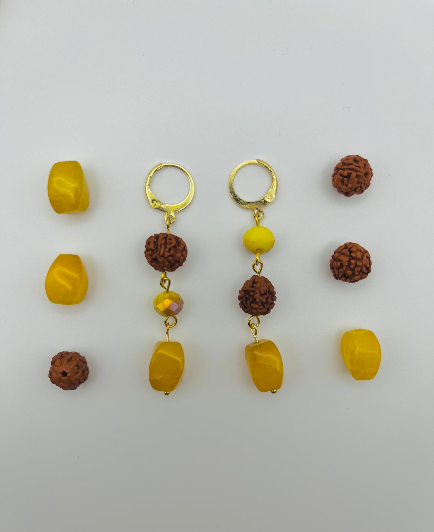 Pendientes Calima – dos aros dorados con cuenta redonda de rudraksha marrón, cuenta facetada amarilla ámbar y cuenta grande ovalada de ámbar amarillo natural, con cuentas de rudraksha y ámbar amarillo sueltas, sobre fondo blanco, hechos a mano en Murcia
