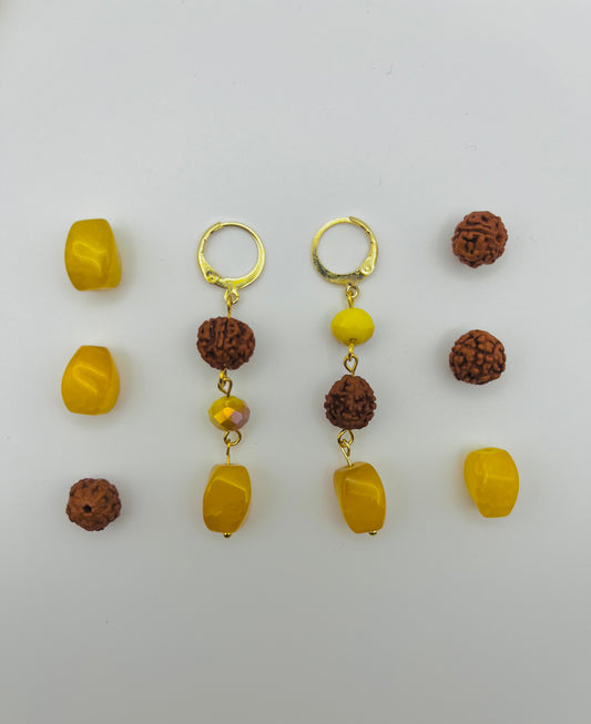 Pendientes Calima – dos aros dorados con cuenta redonda de rudraksha marrón, cuenta facetada amarilla ámbar y cuenta grande ovalada de ámbar amarillo natural, con cuentas de rudraksha y ámbar amarillo sueltas, sobre fondo blanco, hechos a mano en Murcia