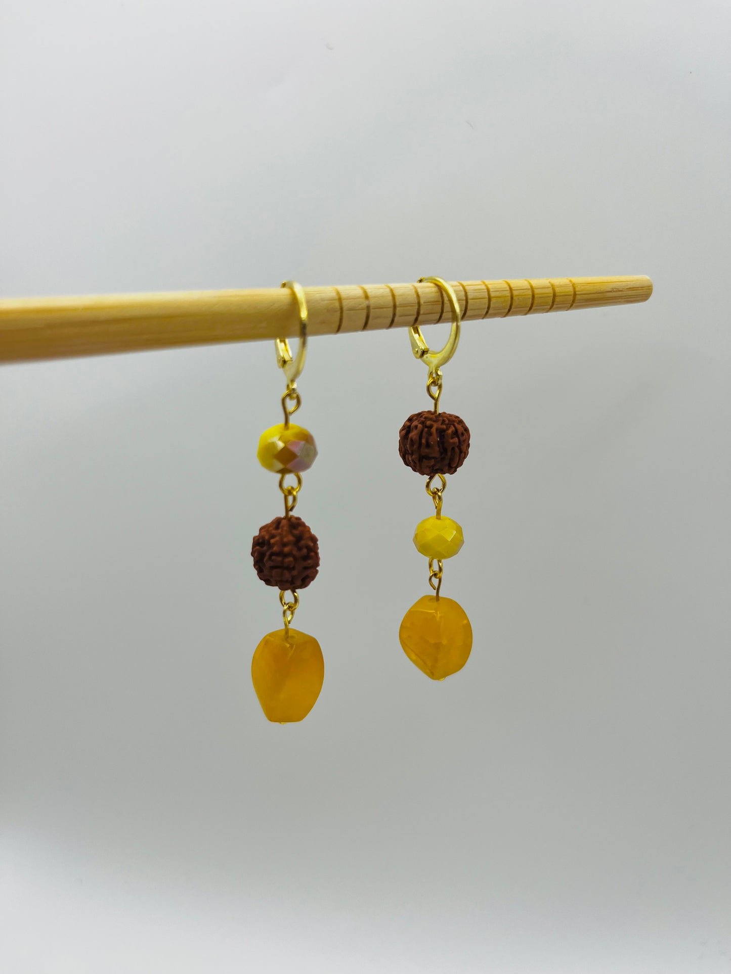 Pendientes Calima – dos aros dorados con cuenta redonda de rudraksha marrón, cuenta facetada ámbar y cuenta grande ovalada de ámbar amarillo natural, colgados de palito de bambú, fondo gris claro, hechos a mano en Murcia