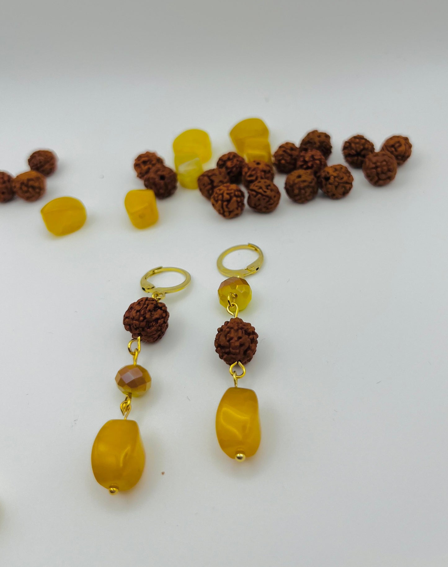 Pendientes Calima – dos aros dorados con cuenta redonda de rudraksha marrón, cuenta facetada ámbar y cuenta grande ovalada de ámbar amarillo natural, en primer plano sobre fondo blanco, con montón de cuentas de rudraksha y ámbar amarillo desenfocadas al fondo, hechos a mano en Murcia