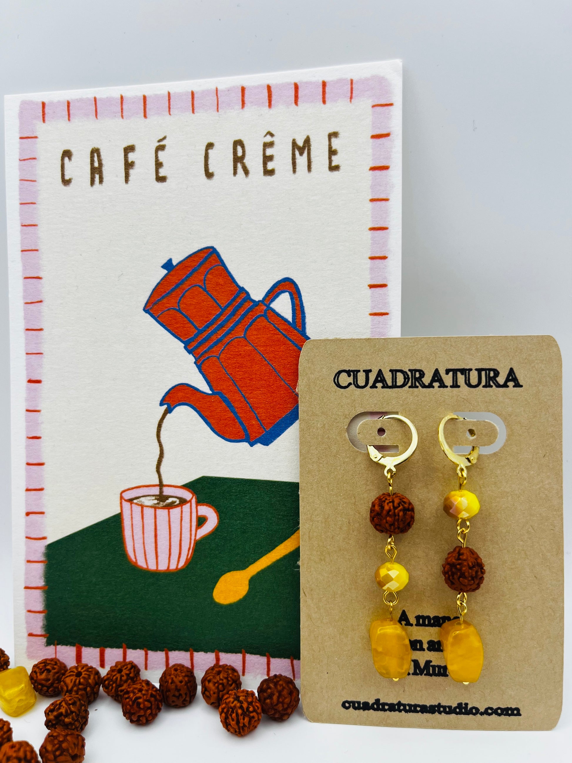 Pendientes Calima – pendientes simétricos con aro dorado, cuenta redonda de rudraksha marrón, cuenta facetada ámbar y cuenta grande ovalada de ámbar amarillo natural, en tarjeta Cuadratura apoyada en postal ilustrada Café Crème con cafetera naranja, con cuentas de rudraksha y ámbar amarillo sueltas en la base, fondo blanco, hechos a mano en Murcia