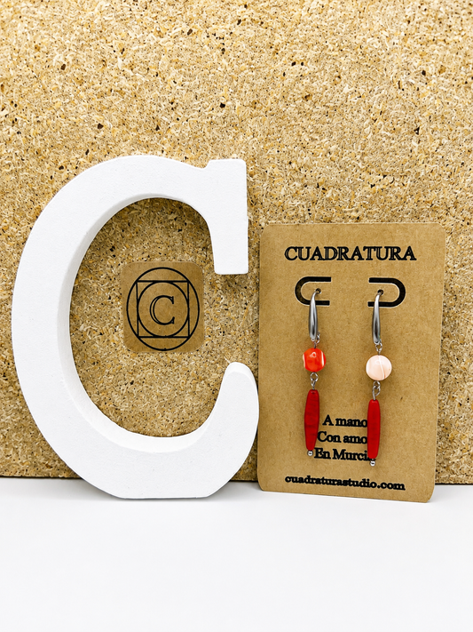 Pendientes asimétricos de Cuadratura Studio en tarjeta kraft: aro de acero inoxidable plateado con cuenta de howlita naranja y barra de coral rojo, y aro con cuenta de howlita rosa y barra de coral rojo. Montados a mano en Murcia.`