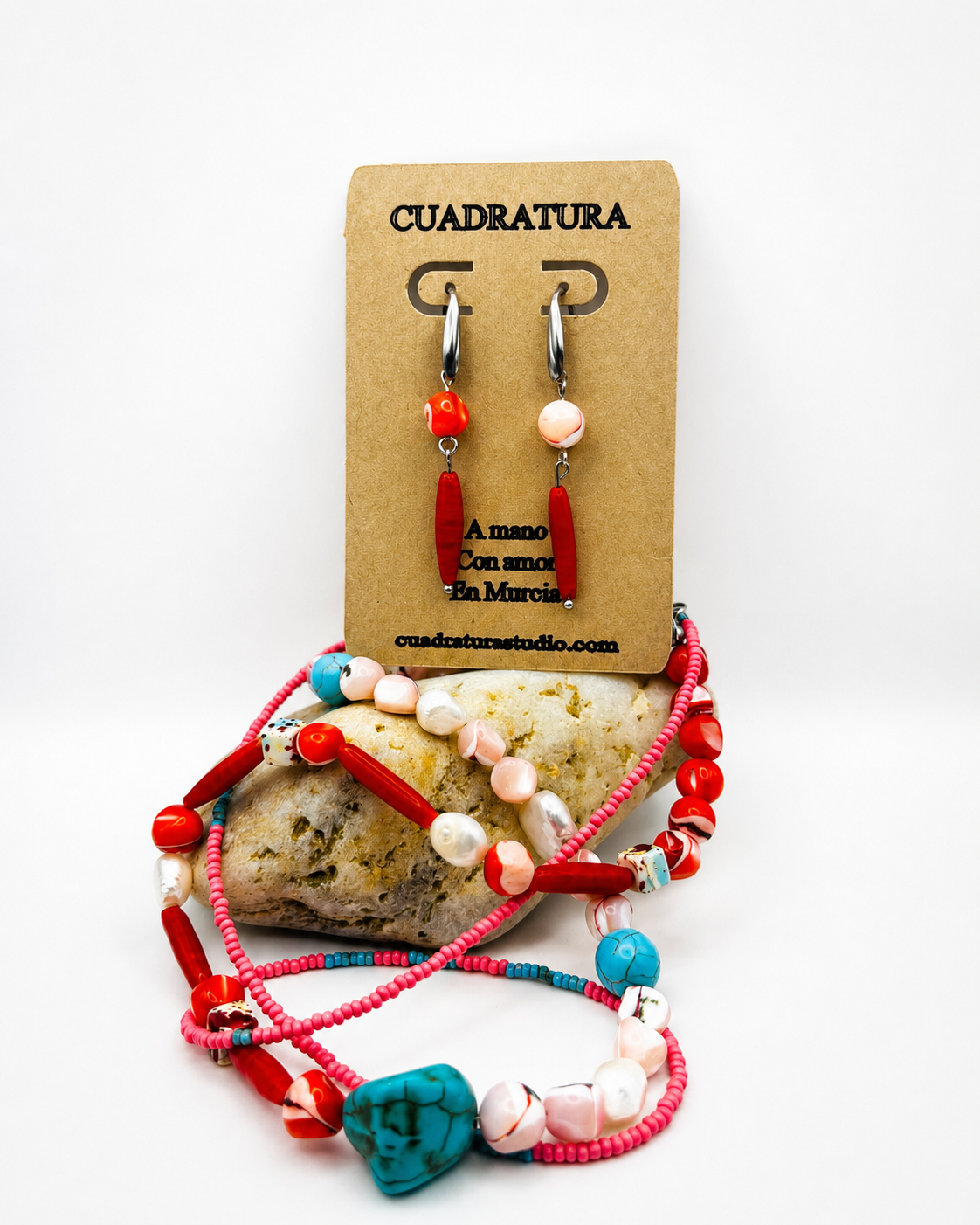 Pendientes de Cuadratura Studio en tarjeta kraft con aro plateado, cuenta de coral naranja y barra de coral rojo, junto a collar y pulsera de semillas rosas, coral rojo, howlita turquesa y nácar, sobre piedra natural. Montados a mano en Murcia.`