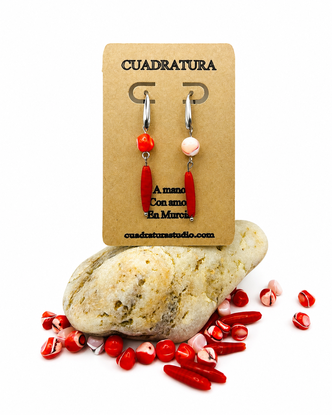 Pendientes asimétricos de Cuadratura Studio en tarjeta kraft: aro plateado con cuenta de coral rojo y barra de coral rojo, y aro con cuenta de howlita rosa y barra de coral rojo, sobre piedra natural rodeada de cuentas de coral sueltas. Montados a mano en Murcia.`