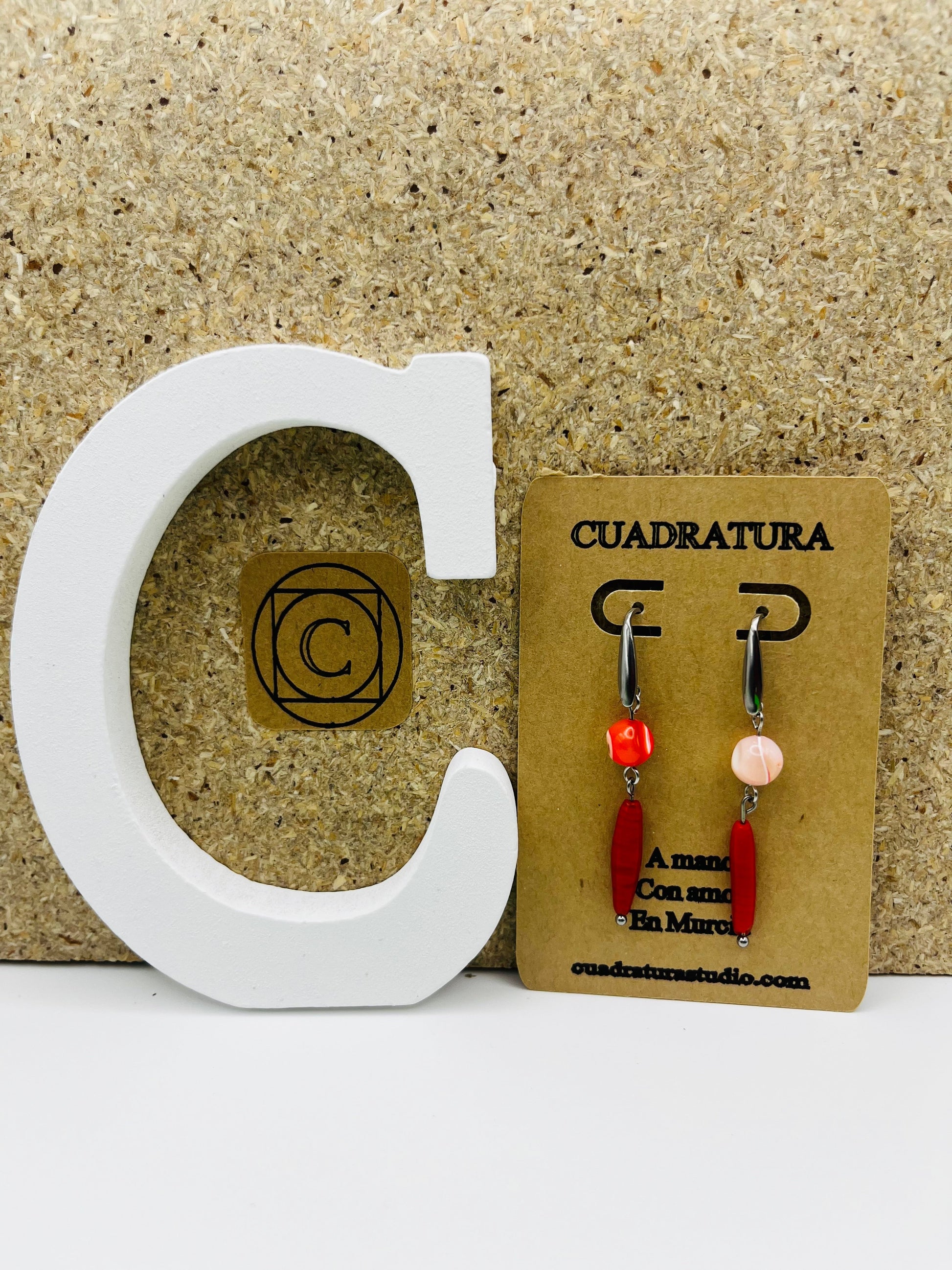 Pendientes Coral – pendientes asimétricos con gancho plateado, cuenta redonda de howlita roja y cuenta ovalada alargada de howlita roja en un pendiente, y cuenta redonda de howlita rosa palo y cuenta ovalada alargada de howlita roja en el otro, en tarjeta Cuadratura apoyada en letra C blanca, fondo de corcho, hechos a mano en Murcia