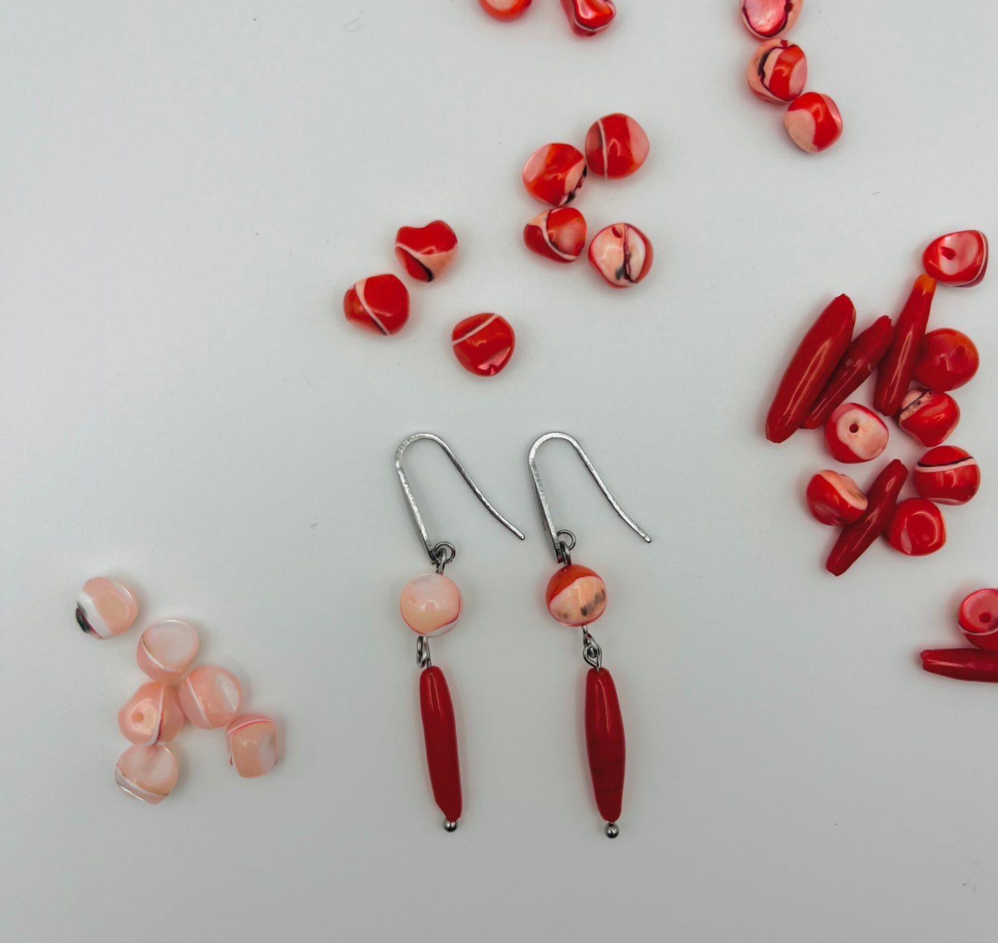 Pendientes Coral – dos ganchos plateados con cuenta redonda de howlita rosa palo con vetas rojas y cuenta ovalada alargada de howlita roja oscura, con cuentas redondas de howlita roja y rosa y cuentas ovaladas alargadas de howlita roja sueltas esparcidas, fondo blanco, hechos a mano en Murcia