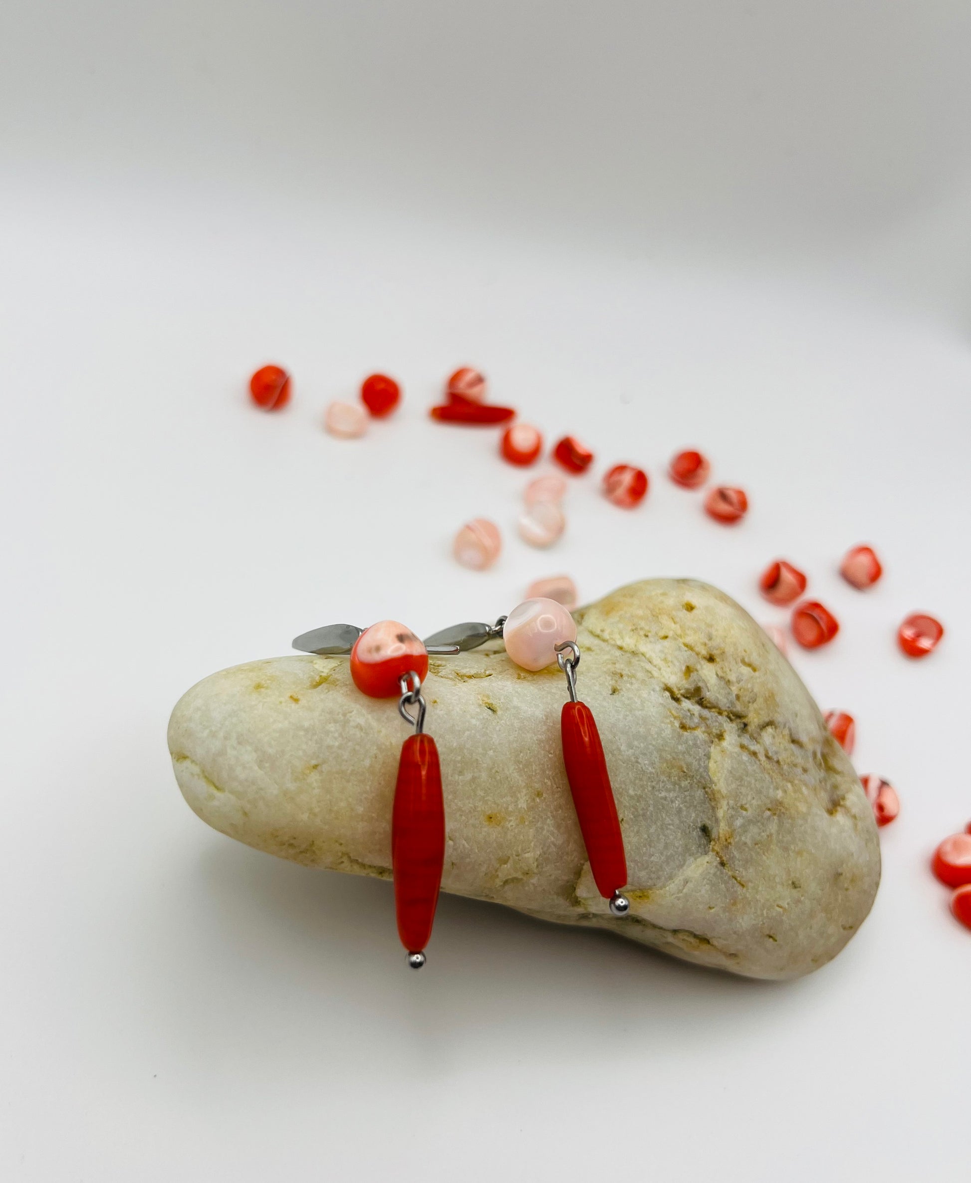 Pendientes Coral – pendientes asimétricos con gancho plateado, cuenta redonda de howlita roja con vetas en un pendiente y cuenta redonda de howlita rosa palo en el otro, ambos con cuenta ovalada alargada de howlita roja oscura colgando, apoyados sobre piedra de río blanca, con cuentas de howlita roja y rosa sueltas al fondo, fondo blanco, hechos a mano en Murcia