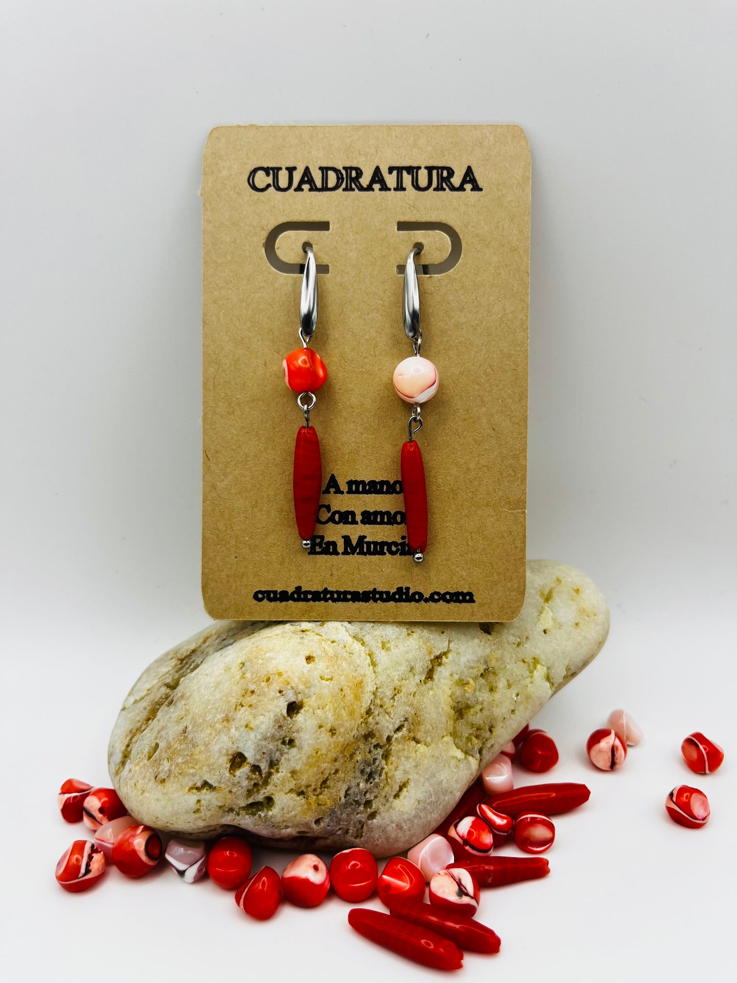Pendientes Coral – pendientes asimétricos con aro plateado, cuenta redonda de howlita roja con vetas en un pendiente y cuenta redonda de howlita rosa palo en el otro, ambos con cuenta ovalada alargada de howlita roja oscura colgando, en tarjeta Cuadratura apoyada sobre piedra de río blanca, con cuentas de howlita roja y rosa sueltas alrededor, fondo gris claro, hechos a mano en Murcia

