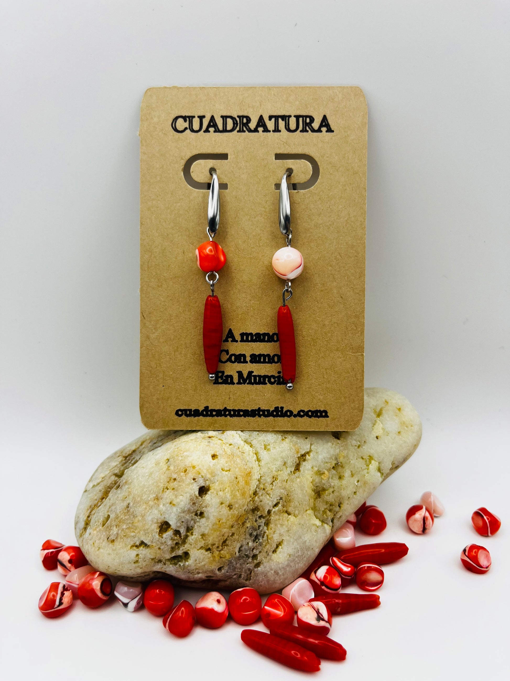 Pendientes Coral – pendientes asimétricos con aro plateado, cuenta redonda de howlita roja con vetas en un pendiente y cuenta redonda de howlita rosa palo en el otro, ambos con cuenta ovalada alargada de howlita roja oscura colgando, en tarjeta Cuadratura apoyada sobre piedra de río blanca, con cuentas de howlita roja y rosa sueltas alrededor, fondo gris claro, hechos a mano en Murcia

