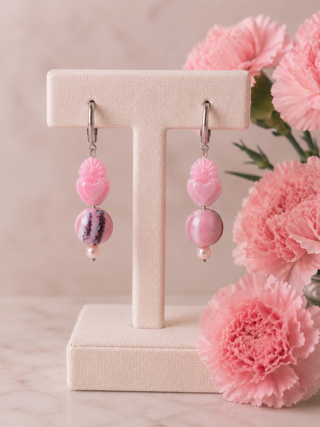 Pendientes artesanales Cuore sobre expositor de joyería. Diseño asimétrico con aros de acero inoxidable plateado, corazones tallados rosa, una cuenta redonda veteada y otra rosa nacarada, rematados con perlas de agua dulce, sobre fondo con claveles rosados.