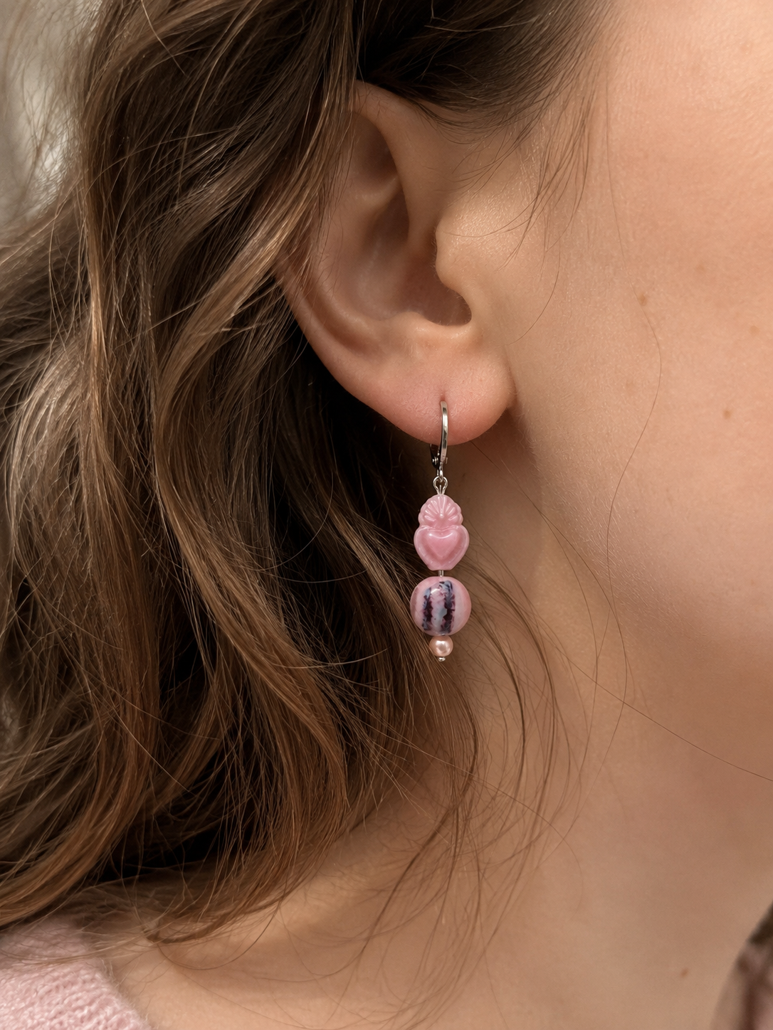 Pendiente artesanal Cuore sobre modelo, con aro de acero inoxidable plateado, corazón tallado rosa, cuenta redonda veteada en rosa, blanco y violeta y perla de agua dulce.