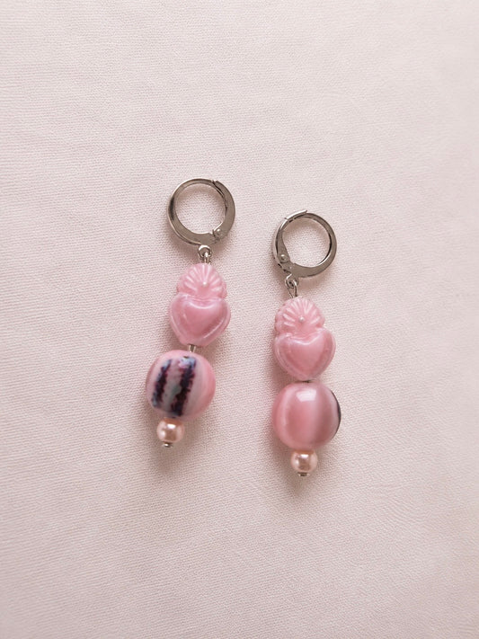 Pendientes artesanales Cuore con aros de acero inoxidable plateado y diseño asimétrico. Ambos incorporan un corazón tallado rosa y una perla de agua dulce; uno lleva una cuenta redonda veteada en rosa, blanco y violeta y el otro una cuenta ovalada rosa nacarada.
