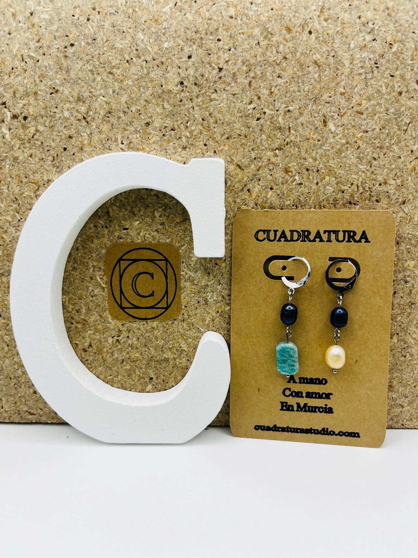 Pendientes Dalia – pendientes asimétricos con aro plateado, cuenta negra de agua dulce, amazonita turquesa facetada y perla barroca amarilla de agua dulce, en tarjeta Cuadratura, sobre letra C blanca y fondo de corcho, hechos a mano en Murcia