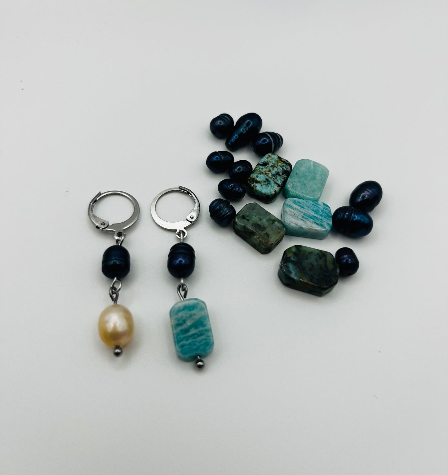 Pendientes Dalia – pendientes asimétricos con aro plateado, perla negra de agua dulce, perla barroca amarilla y amazonita turquesa facetada, con perlas negras y cuentas de amazonita sueltas esparcidas, sobre fondo blanco, hechos a mano en Murcia