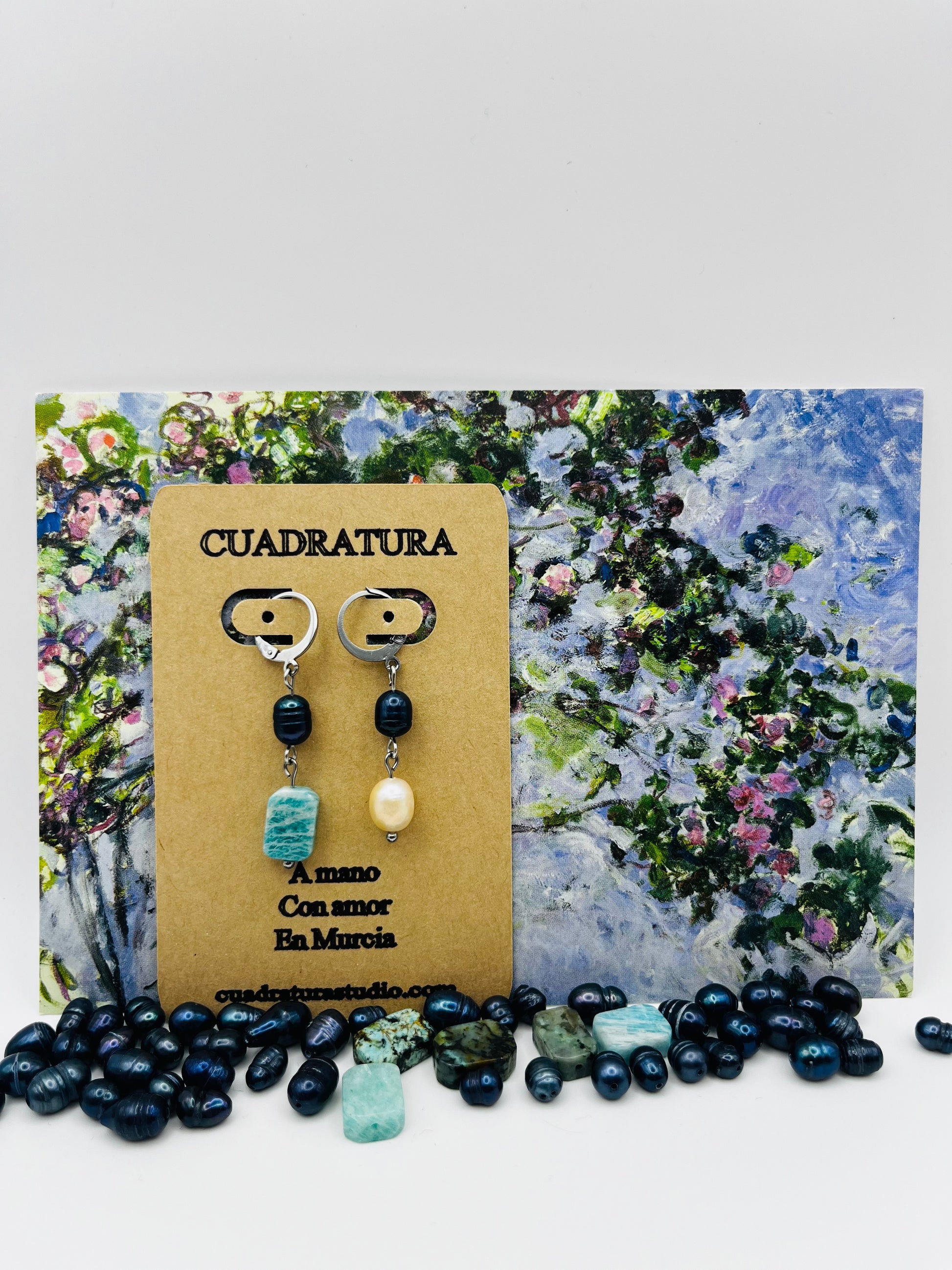 Pendientes Dalia – pendientes asimétricos con aro plateado, perla negra de agua dulce, amazonita turquesa facetada y perla barroca amarilla, en tarjeta Cuadratura, sobre postal de pintura impresionista de jardín con perlas negras y cuentas de amazonita sueltas en la base, hechos a mano en Murcia